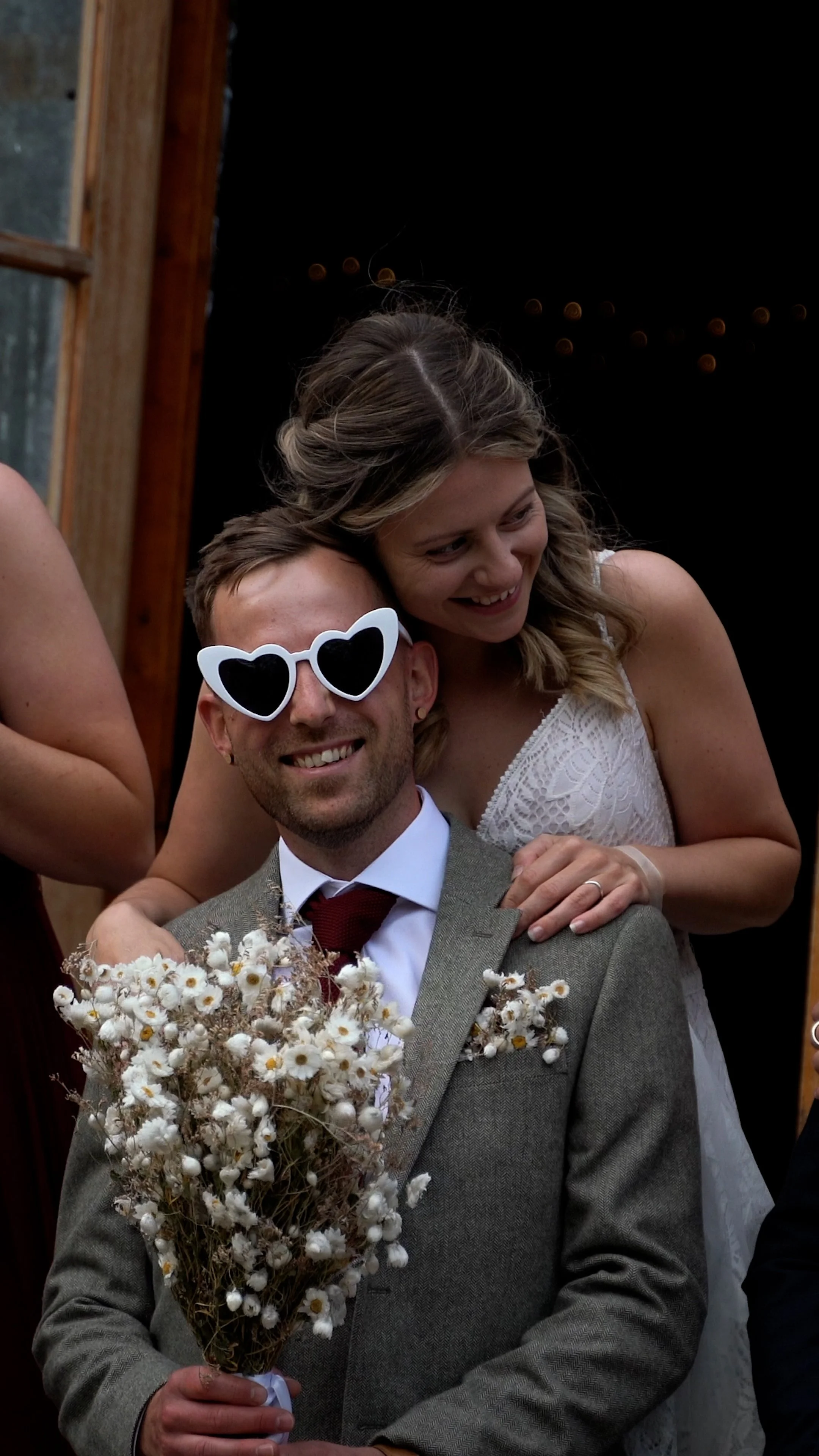 A smiling man wearing heart-shaped sunglasses and a suit holding a bouquet of daisies, with a woman leaning over his shoulder smiling, at a wedding or special celebration.