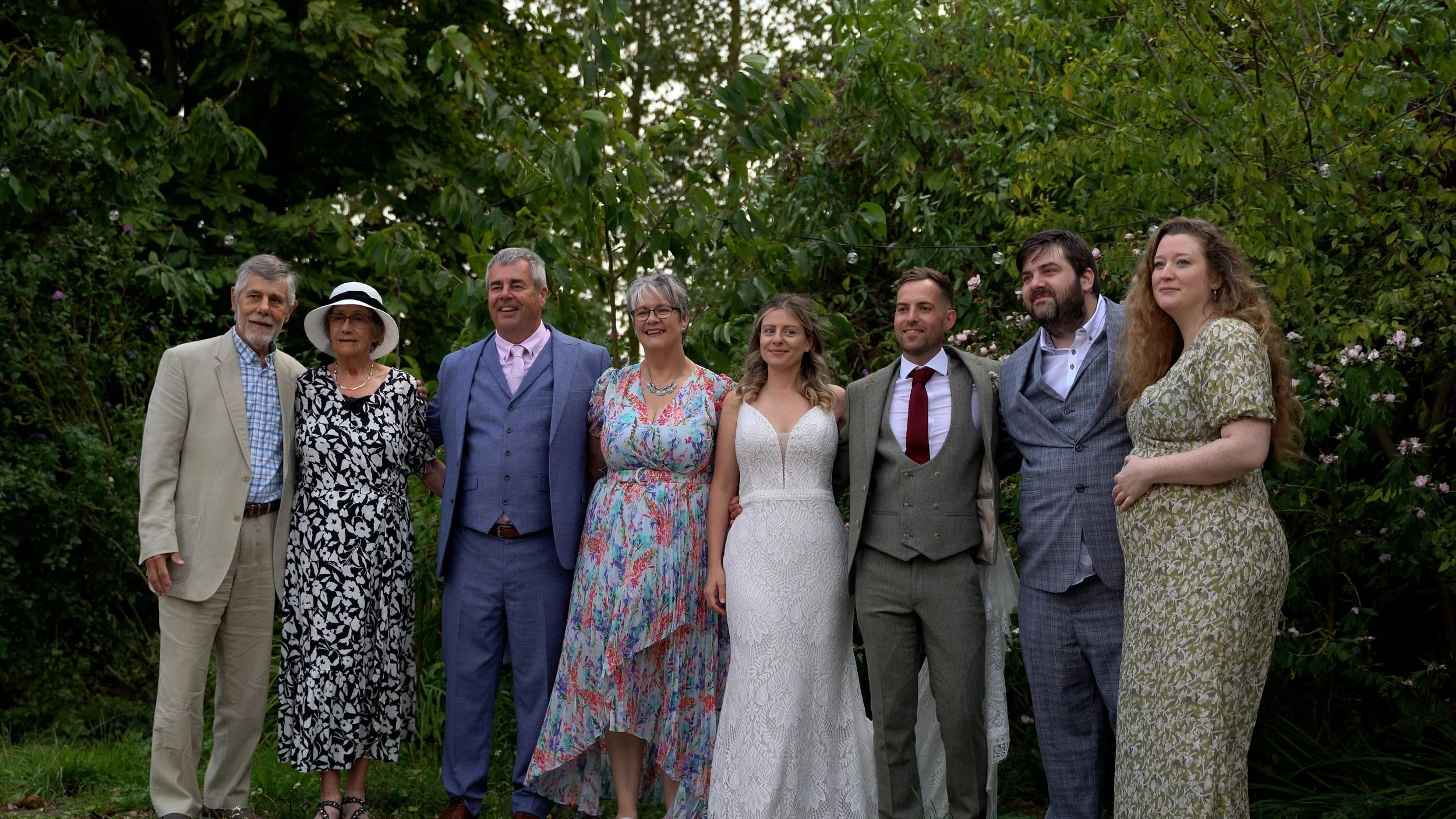 Group of eight smiling people at a wedding in a garden, including a bride in a white lace dress, a groom in a gray suit, and six older adults dressed in formal and semi-formal attire, standing under greenery decorated with string lights.