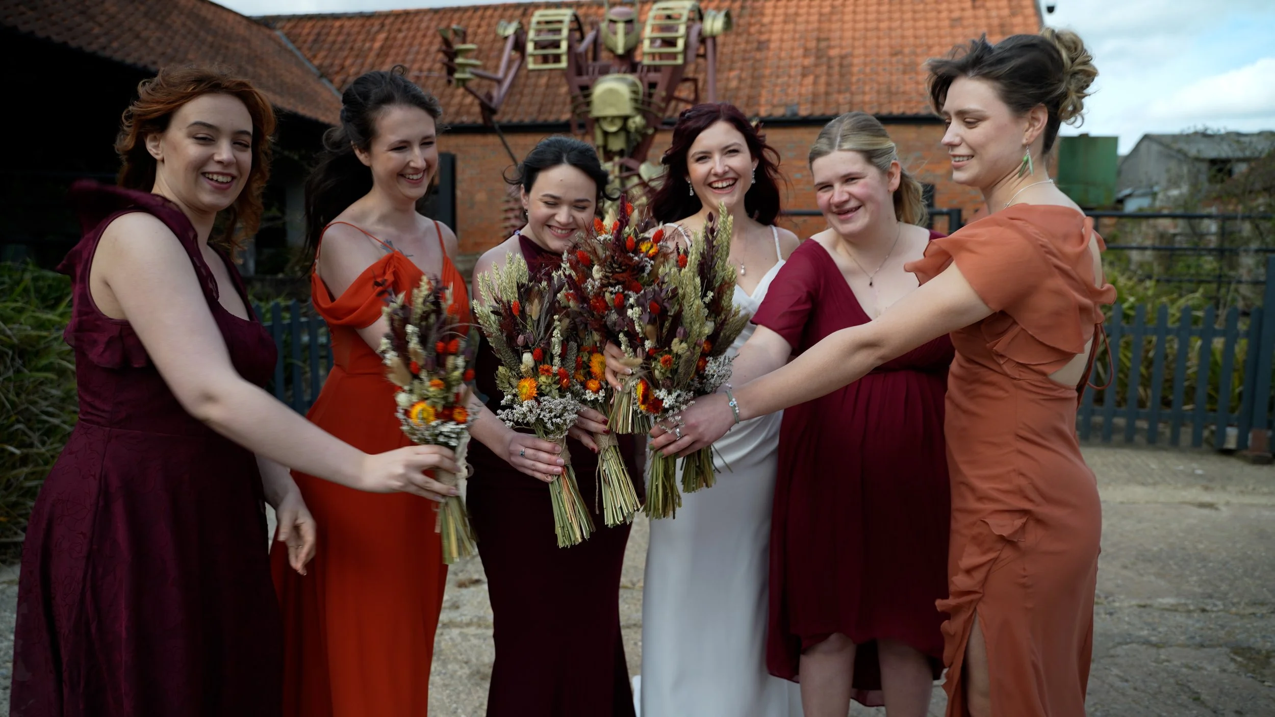 Six women dressed in colorful dresses standing outdoors, holding bouquets of flowers and smiling.