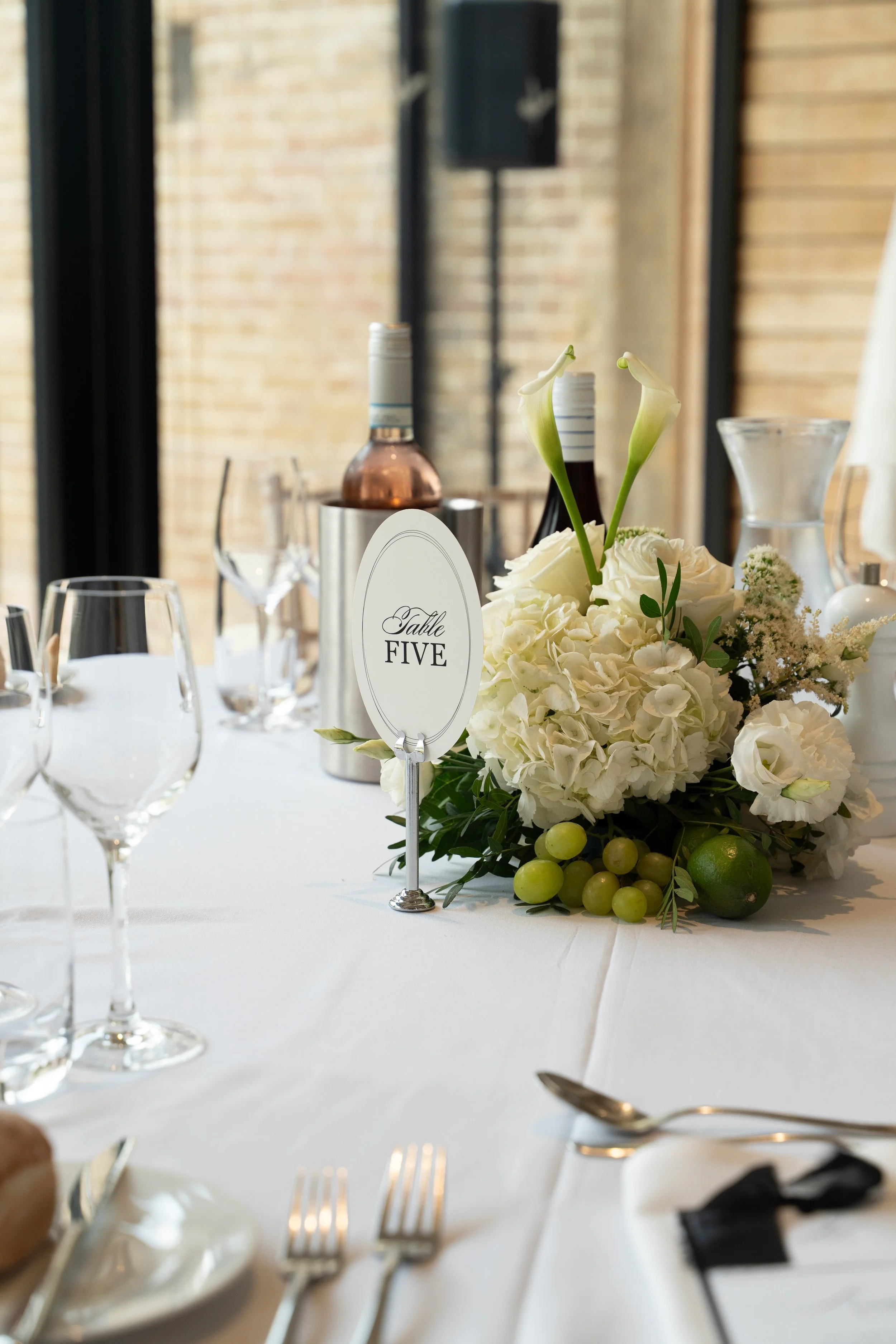 A close-up of a banquet table with white tablecloth, floral centerpiece with white roses, calla lilies, grapes, and lime, wine bottles, glassware, and a table number sign reading 'Table Five' in a bright venue with large windows and exposed brick walls.
