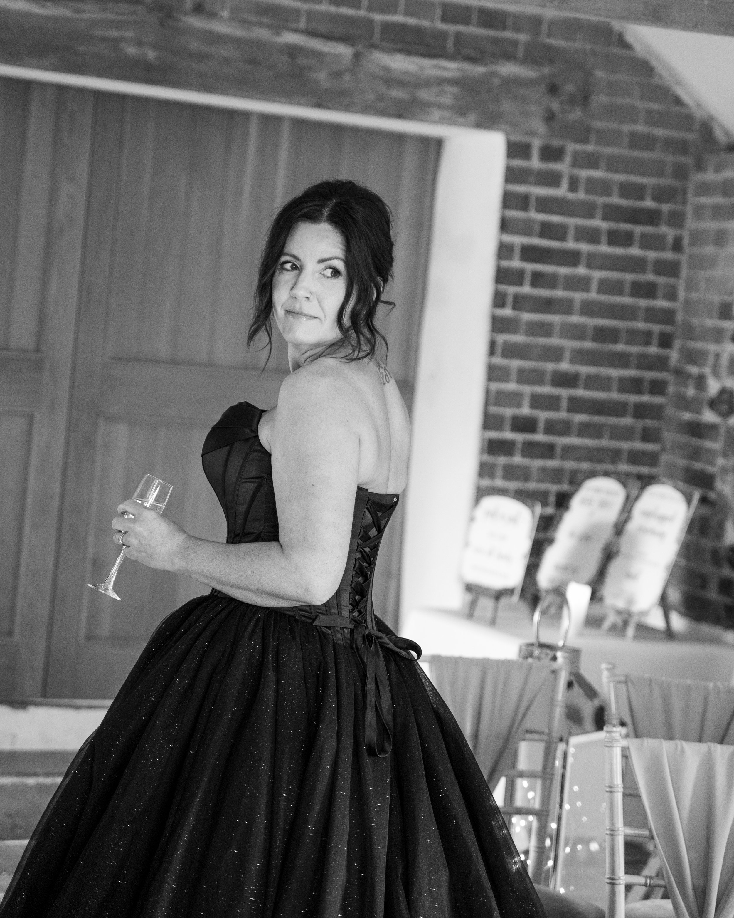 A woman in an elegant strapless black dress with a full tulle skirt holding a champagne flute. She has dark hair styled in loose waves and is standing in a room with wooden walls, brick walls, and chairs.