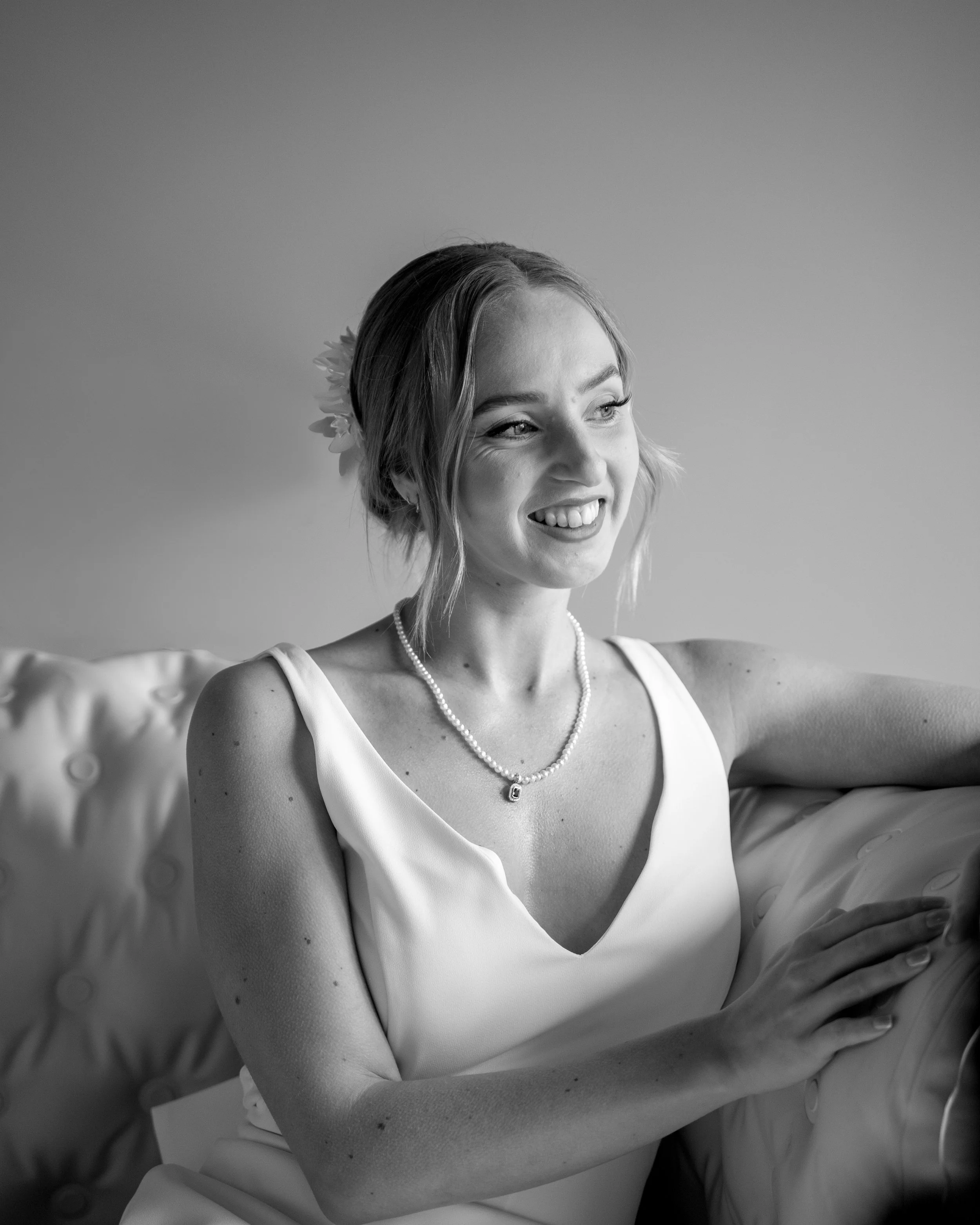 Black and white photo of a woman smiling with her arms crossed. She is wearing a sleeveless dress, a pearl necklace, and a floral hair accessory, seated on a cushioned armchair.