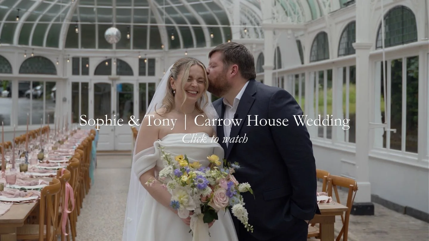 A bride and groom share a happy moment inside a bright, glass-enclosed wedding venue. The bride is holding a colorful bouquet, and the groom is kissing her on the forehead.
