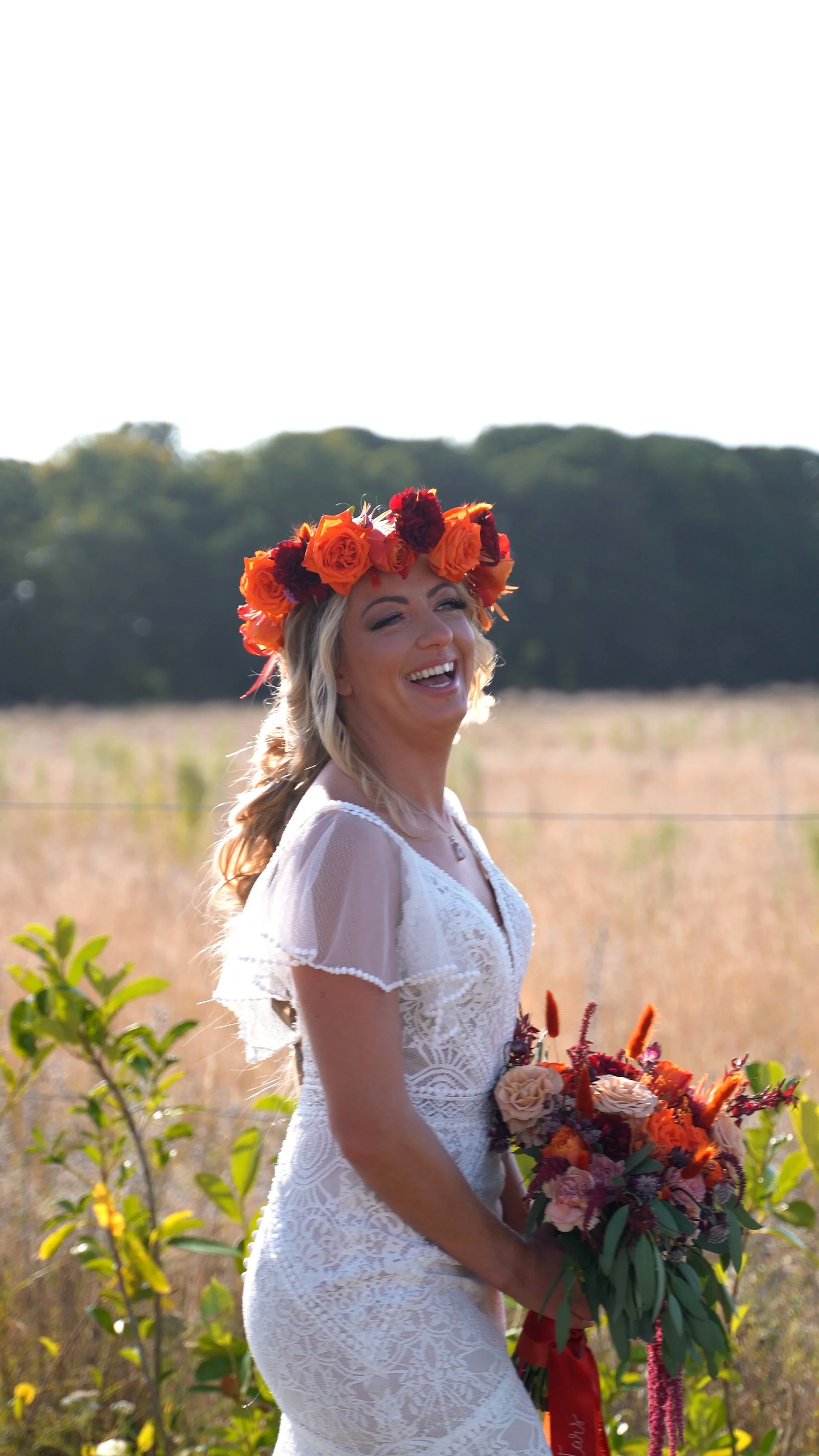 A woman in a white lace dress stands outdoors, smiling with a flower crown of orange and red roses, holding a bouquet of orange and pink flowers, with a field and trees in the background.