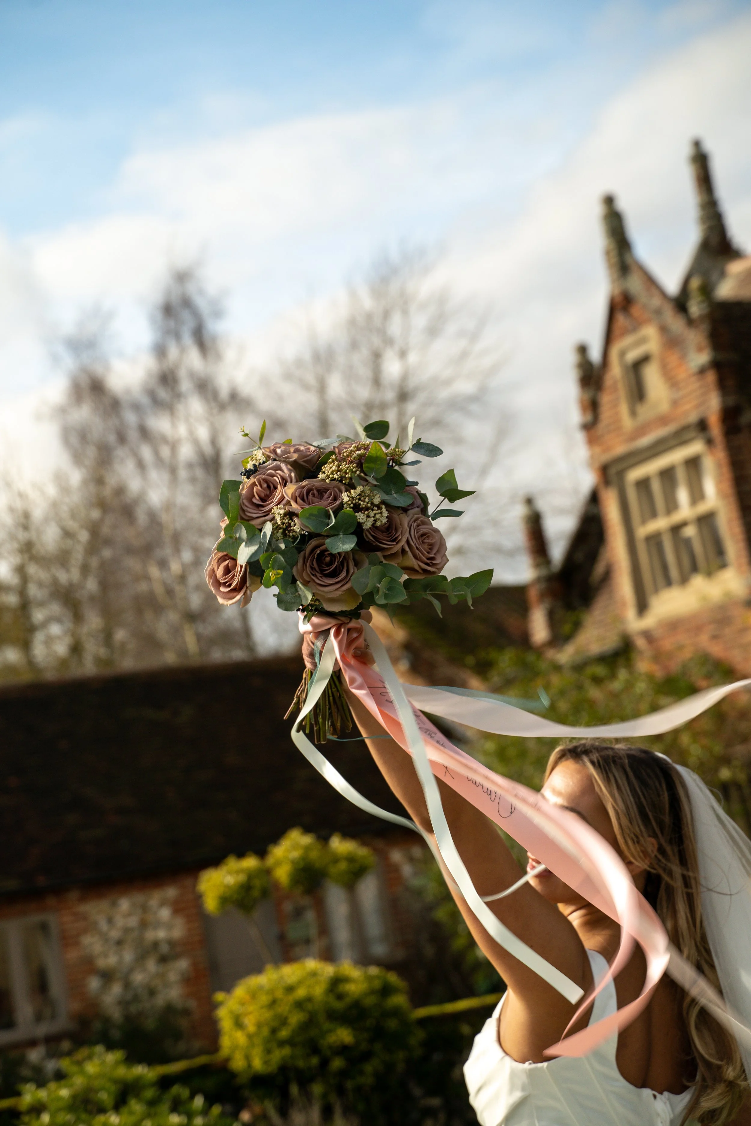 A woman holding a bouquet of flowers and ribbons outdoors, with a historic brick building and trees in the background.
