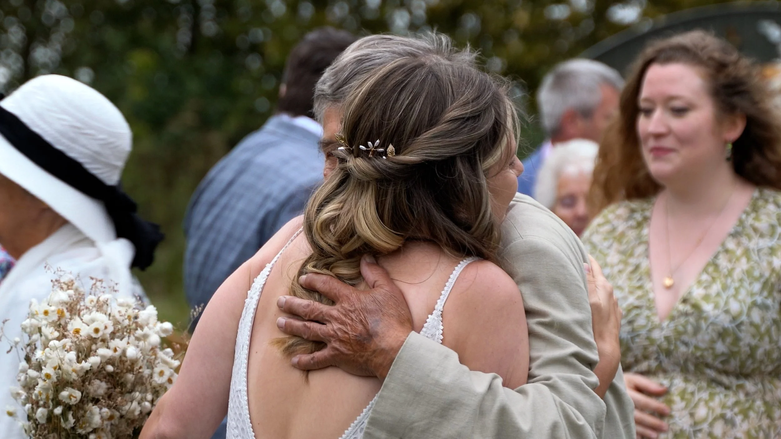 A woman with braided hair embracing an older man at an outdoor gathering, with other people in the background and a bouquet of white flowers in the foreground.