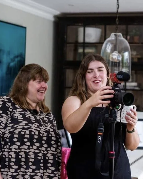 Two women, one older and one younger, are smiling and looking at a camera mounted on a tripod. The younger woman is adjusting the camera in a room with a dark bookshelf and a hanging light fixture overhead.