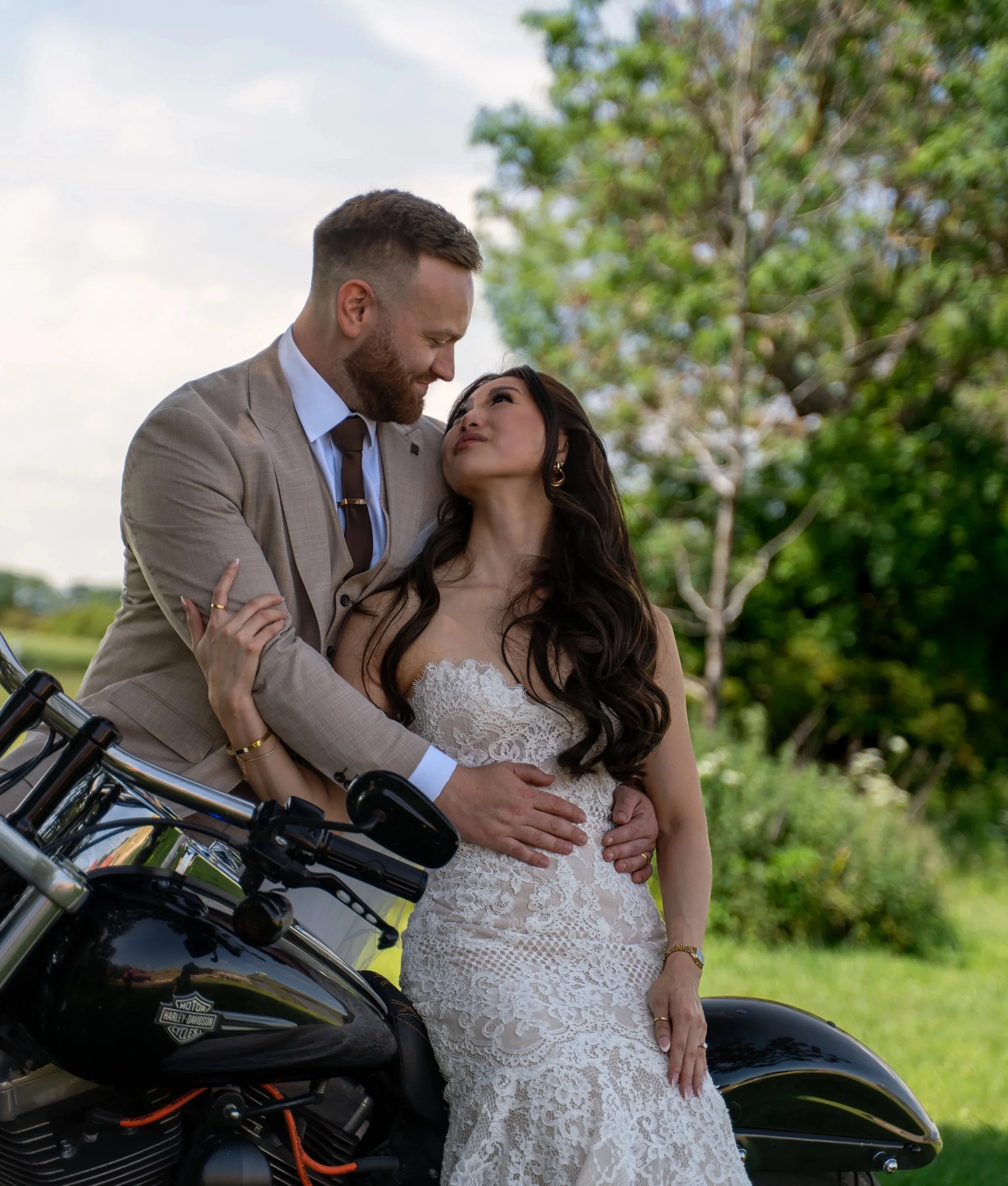 A couple sharing a tender moment outdoors, with the man in a beige suit and the woman in a white lace dress, standing next to a black Harley Davidson motorcycle, surrounded by greenery and trees.