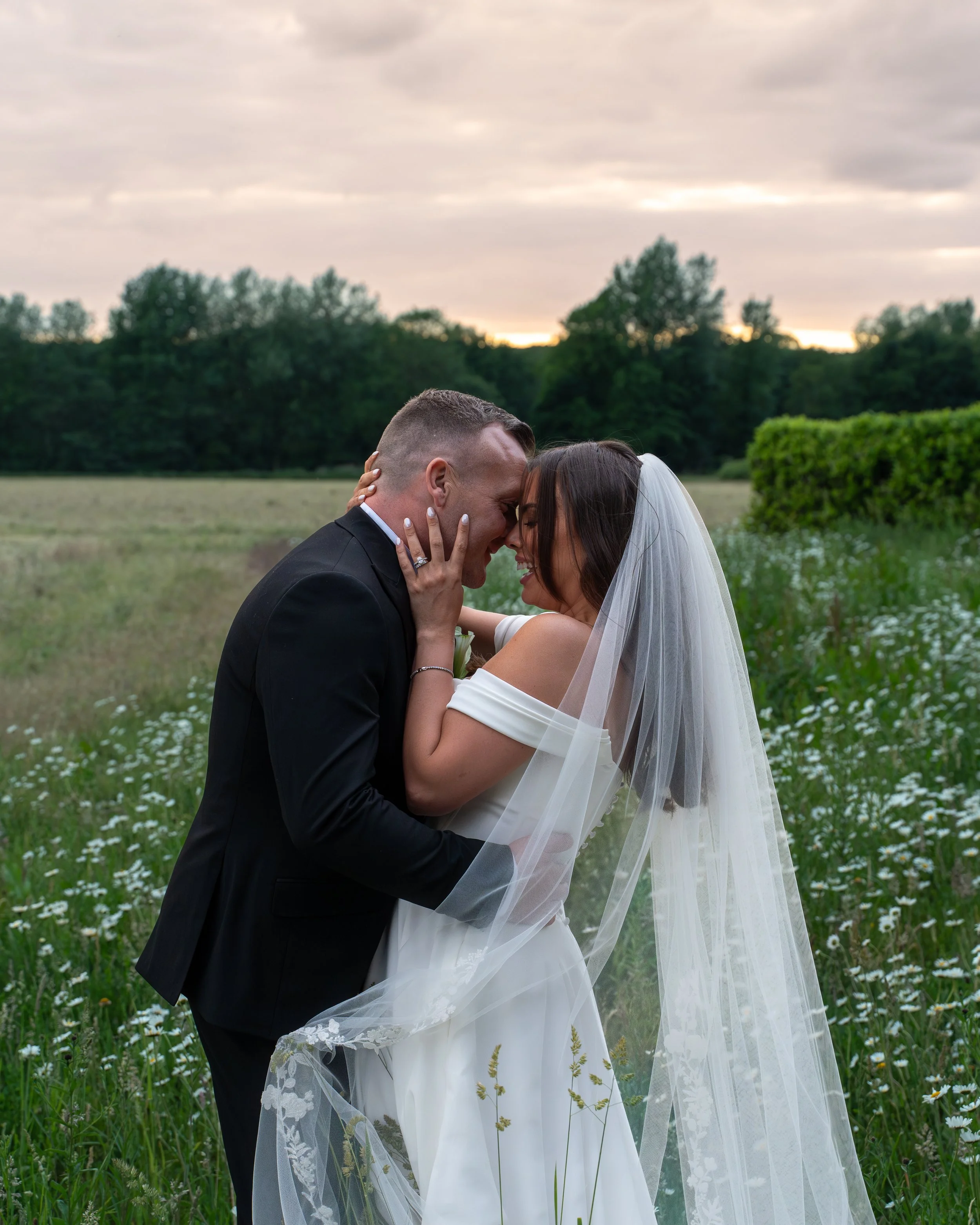 A wedding couple in formal attire sharing an intimate moment outdoors in a field with wildflowers, trees, and a cloudy sky at sunset.