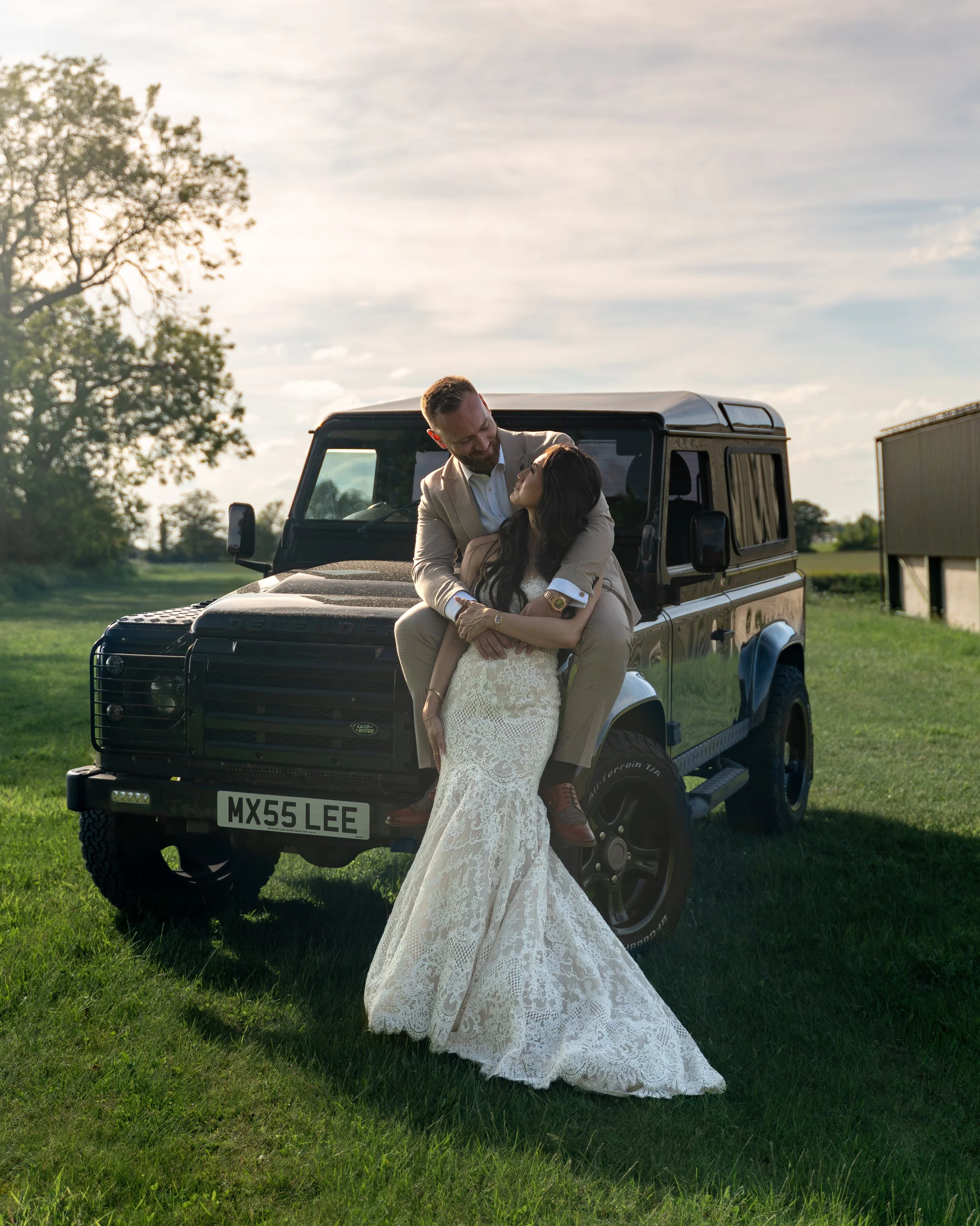 A man and woman are embracing in front of a black off-road vehicle on a grassy field during daytime. The woman is wearing a white lace wedding dress, and the man is dressed in a beige suit. They are looking at each other affectionately.