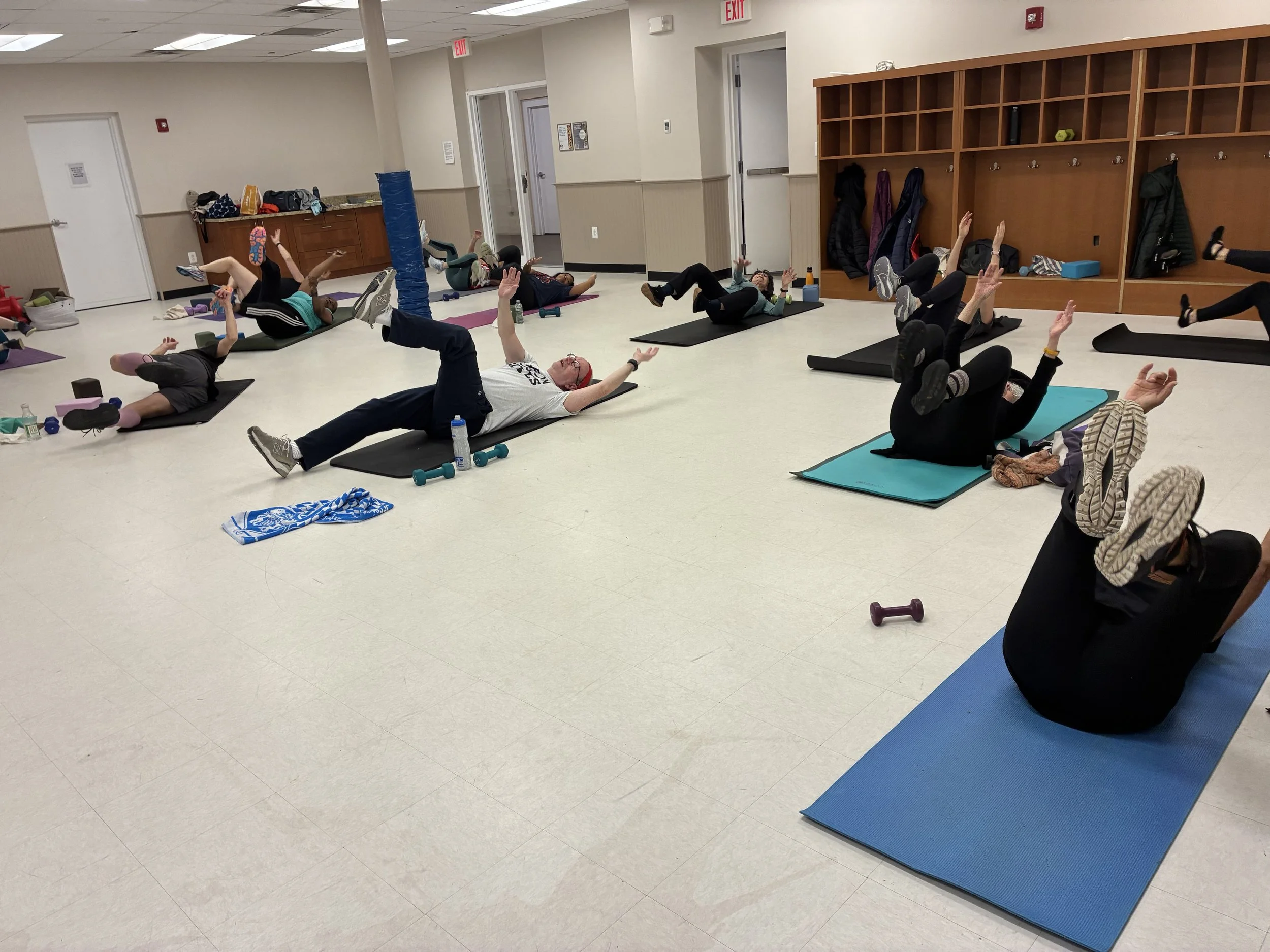 Group of people participating in an indoor fitness class lying on yoga mats and raising their arms and legs, with personal belongings and water bottles around them.