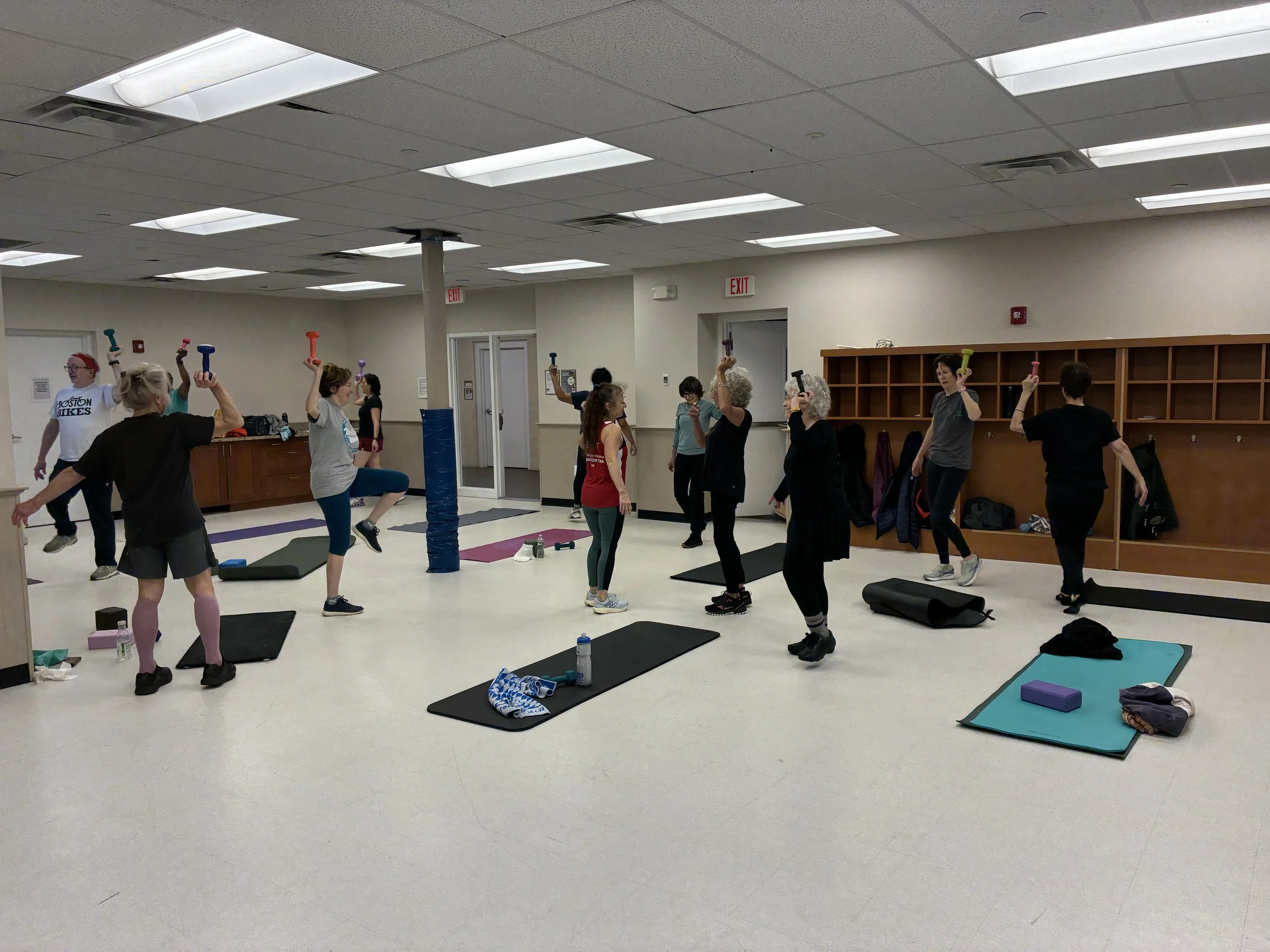 A group of people participating in a fitness class in a large room with bright ceiling lights, yoga mats, and bottles of water on the floor, some holding exercise weights above their heads.