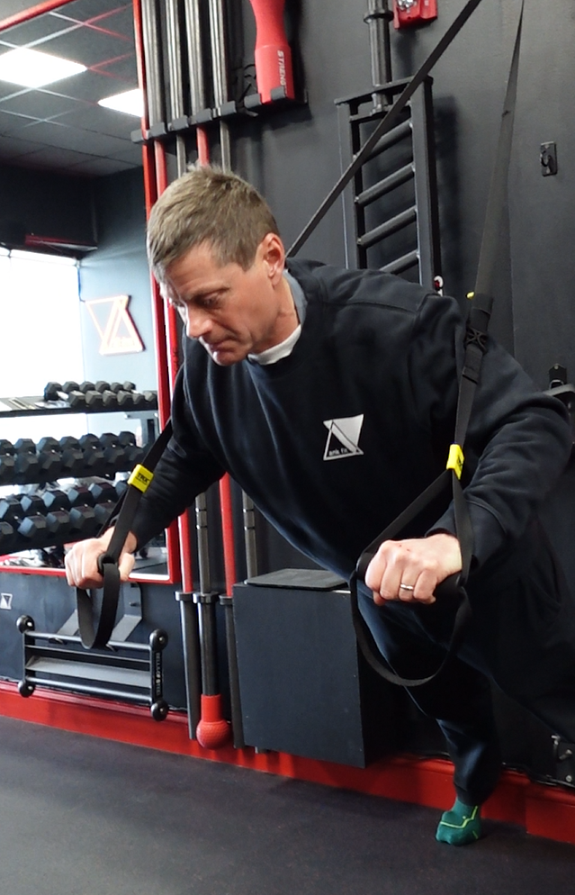 Rob Skinner performing a suspension workout using TRX straps in a gym, with equipment and weights in the background.