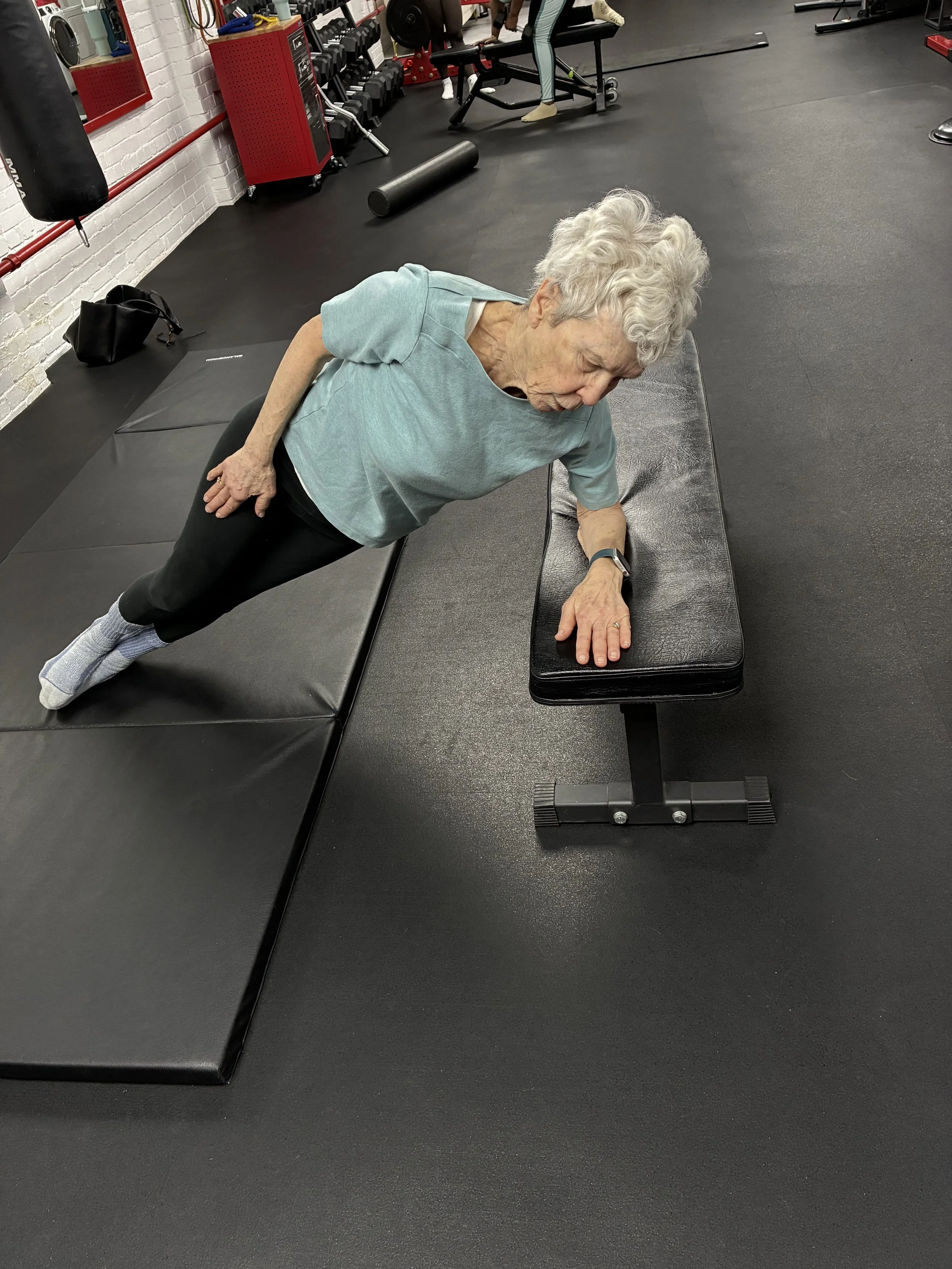 An elderly woman outdoors performing a side plank exercise on a black workout mat, Supportive shoes, and casual clothes, with trees and grass in the background.