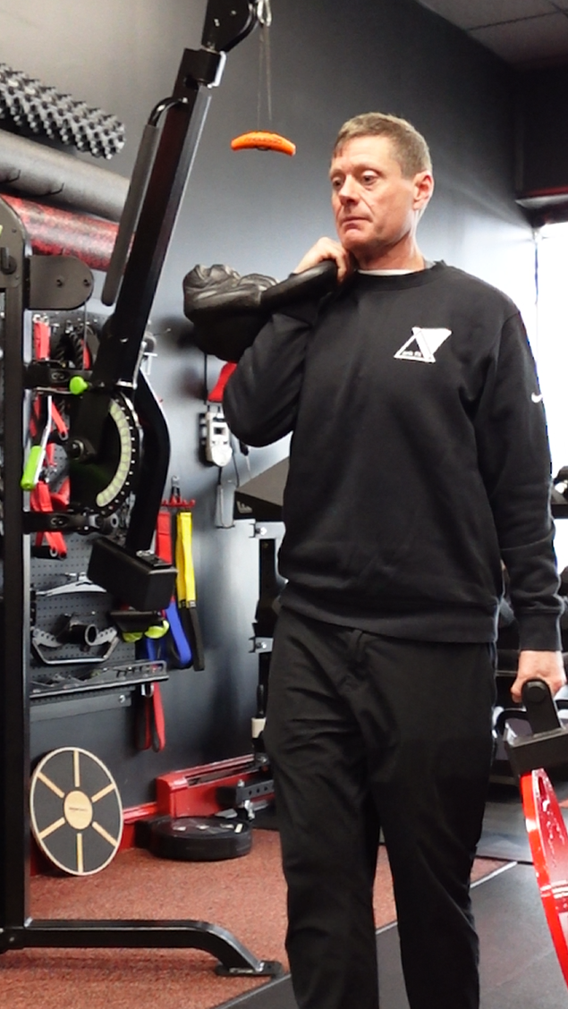 Rob Skinner in black workout clothing lifting a weight on his shoulder in a gym, with exercise equipment and foam rollers in the background.