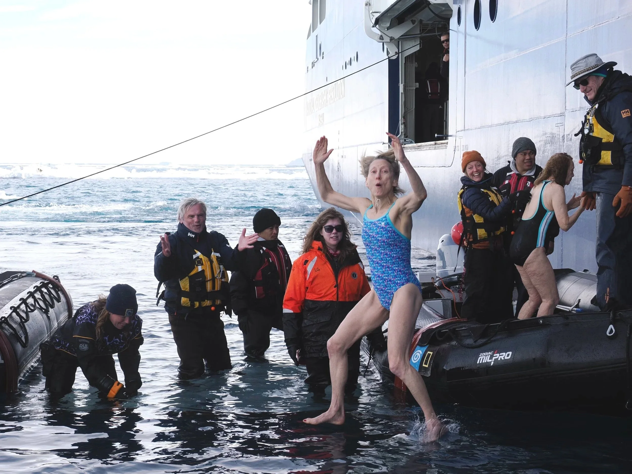 A group of people, some wearing life jackets, are standing in the water next to a large ship. One woman in a swimsuit is stepping onto an inflatable boat, while others are assisting. The scene appears to be an emergency or rescue operation.