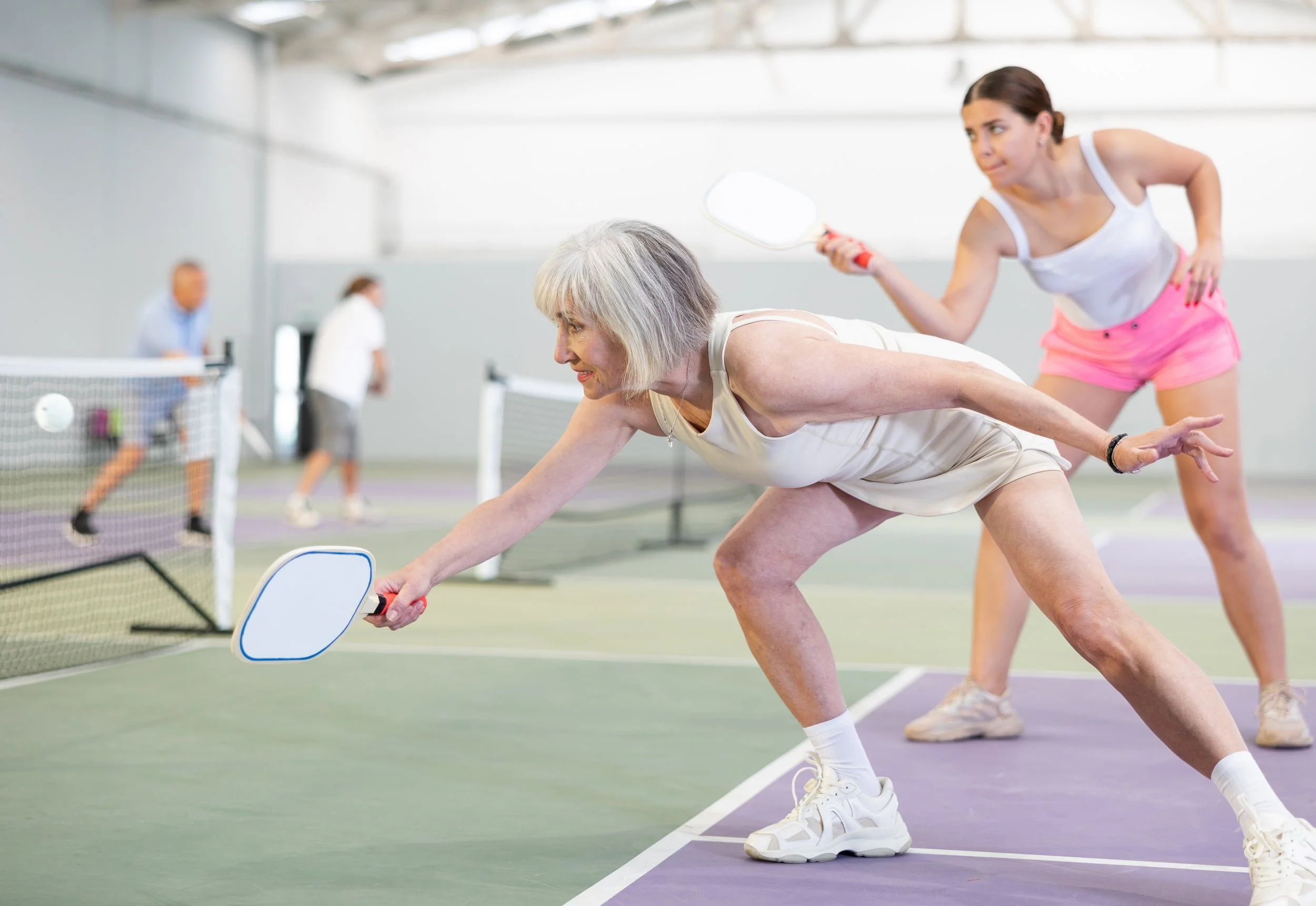 An elderly woman in a white tank top and shorts playing pickleball on an indoor court, with a woman in a white top and pink shorts holding a paddle nearby, and other players in the background.