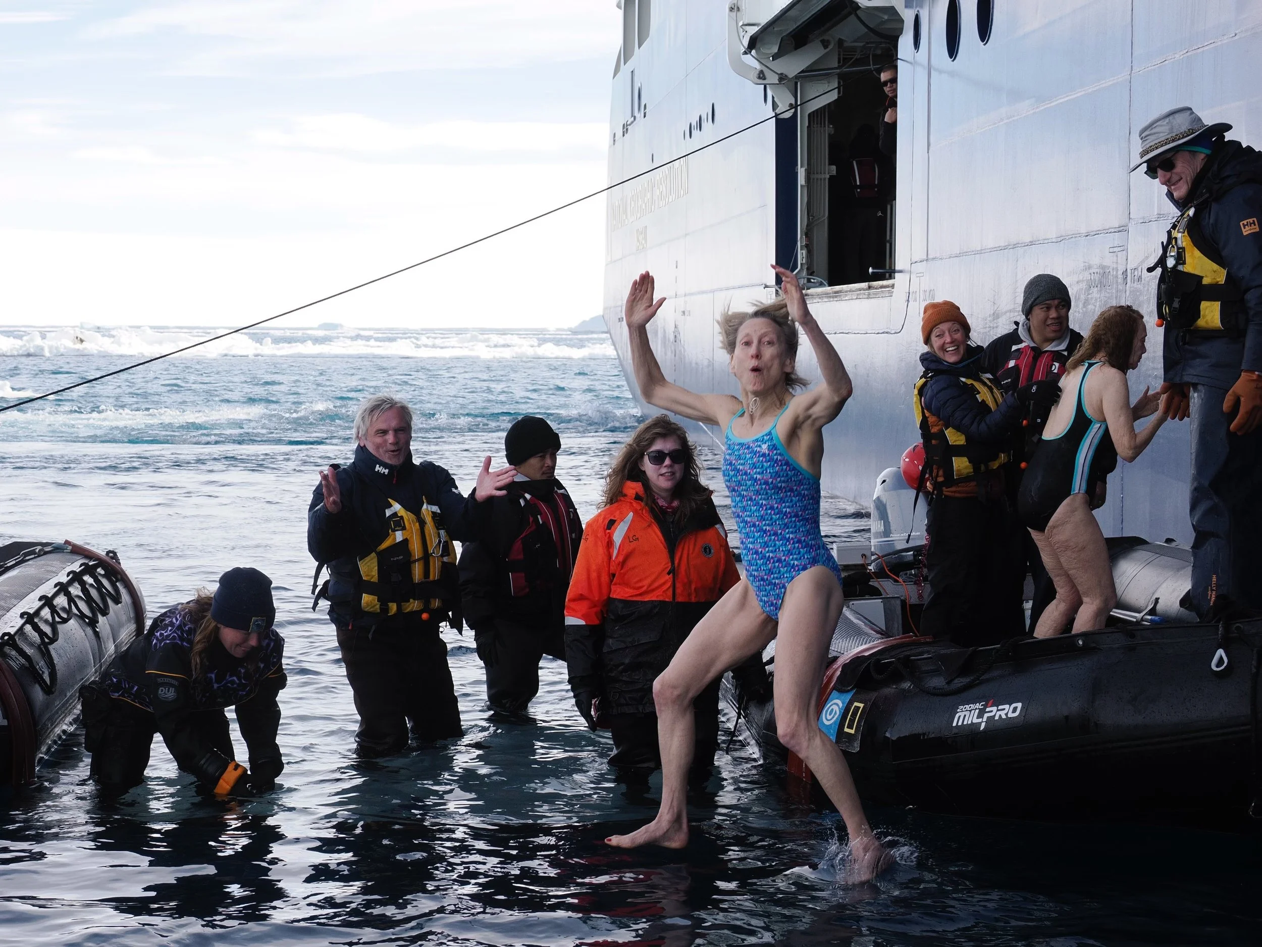 Group of people in water and life jackets preparing to board a boat in icy waters with an iceberg in the background.