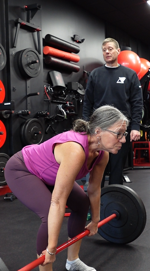 An older woman in a purple workout outfit lifting a barbell in a gym, with a man observing in the background.