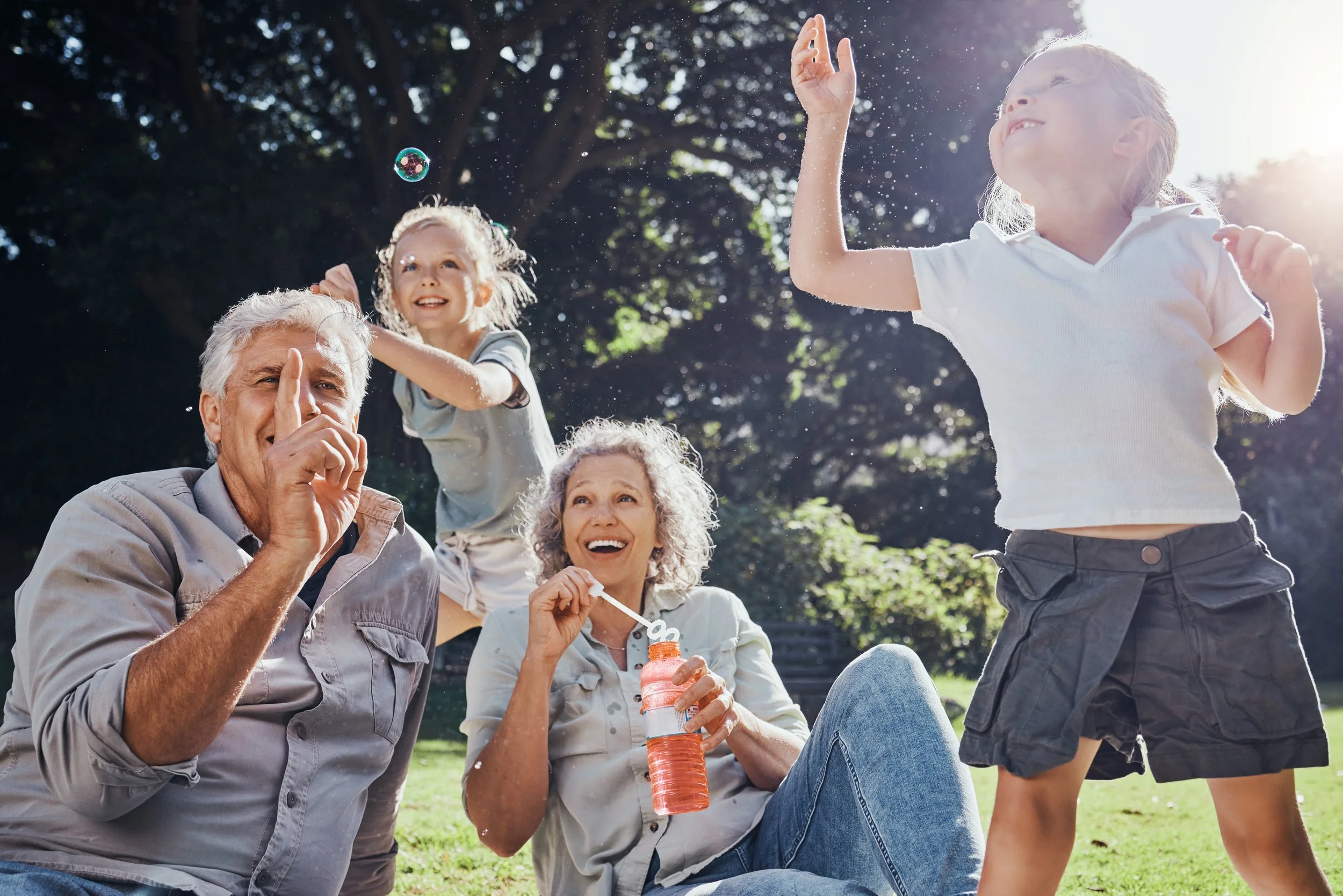 Family outdoors on a sunny day, playing and laughing together with children and grandparents, children blowing bubbles, one grandparent sitting and another standing.