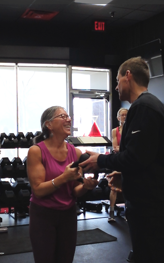 A woman and a man are smiling and shaking hands inside a gym with exercise equipment and large windows in the background.