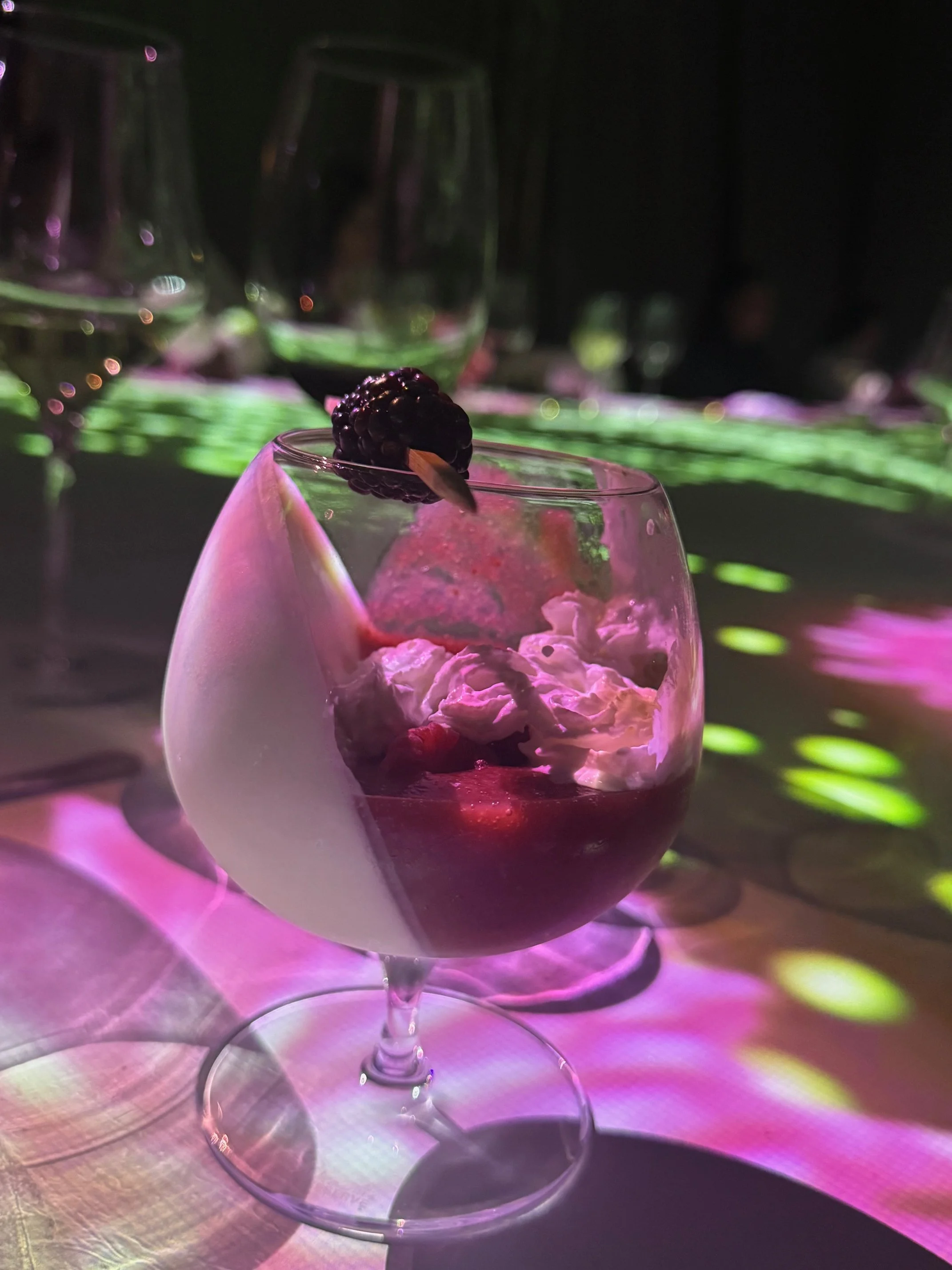 A glass of red wine topped with a blackberry and a small wooden stick, set against a colorful, blurred background with glasses of wine and abstract light reflections.