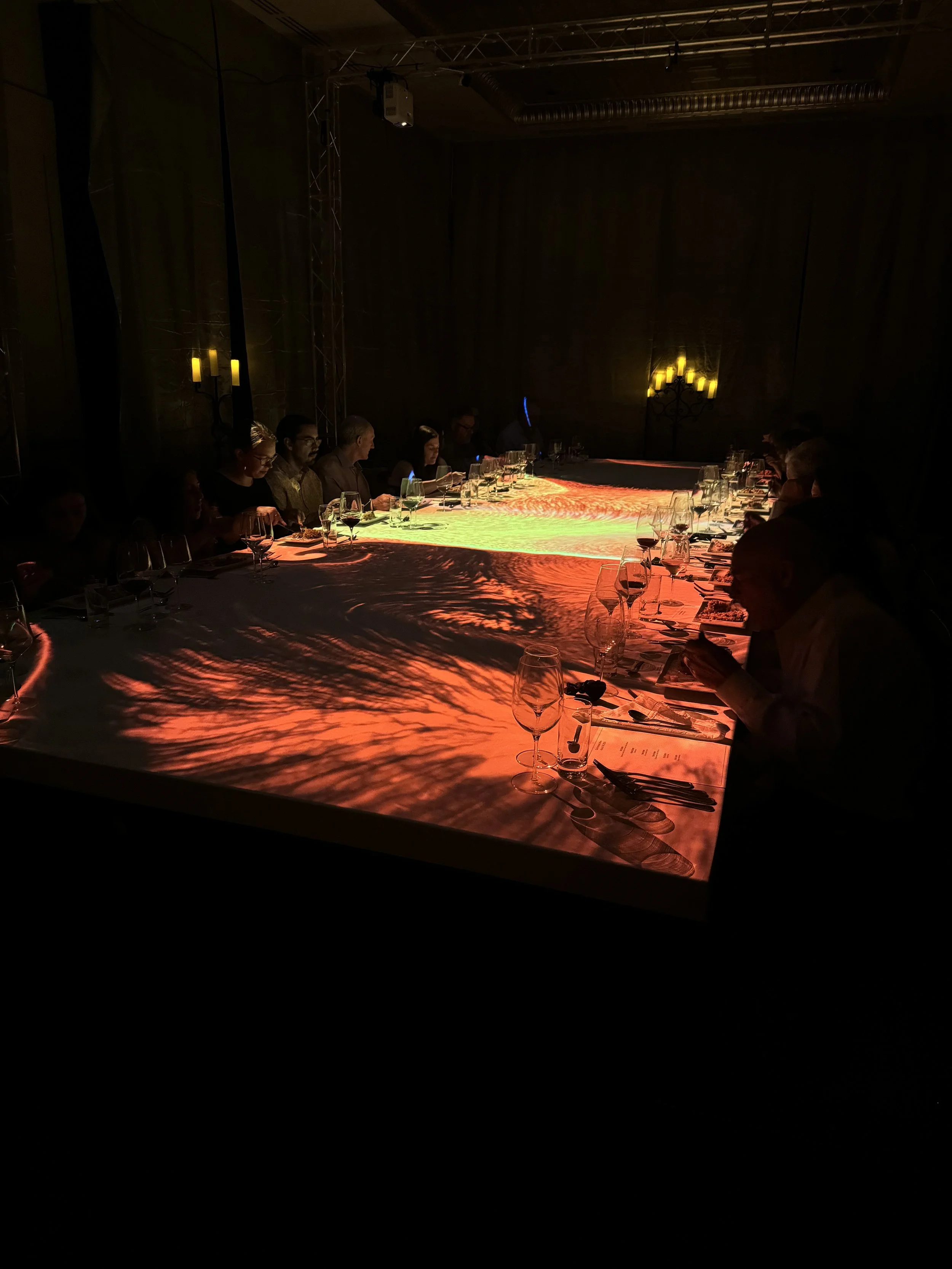People sitting around a long table in a dimly lit room with colorful light projections on the table, candles on the walls, and some people dining quietly.