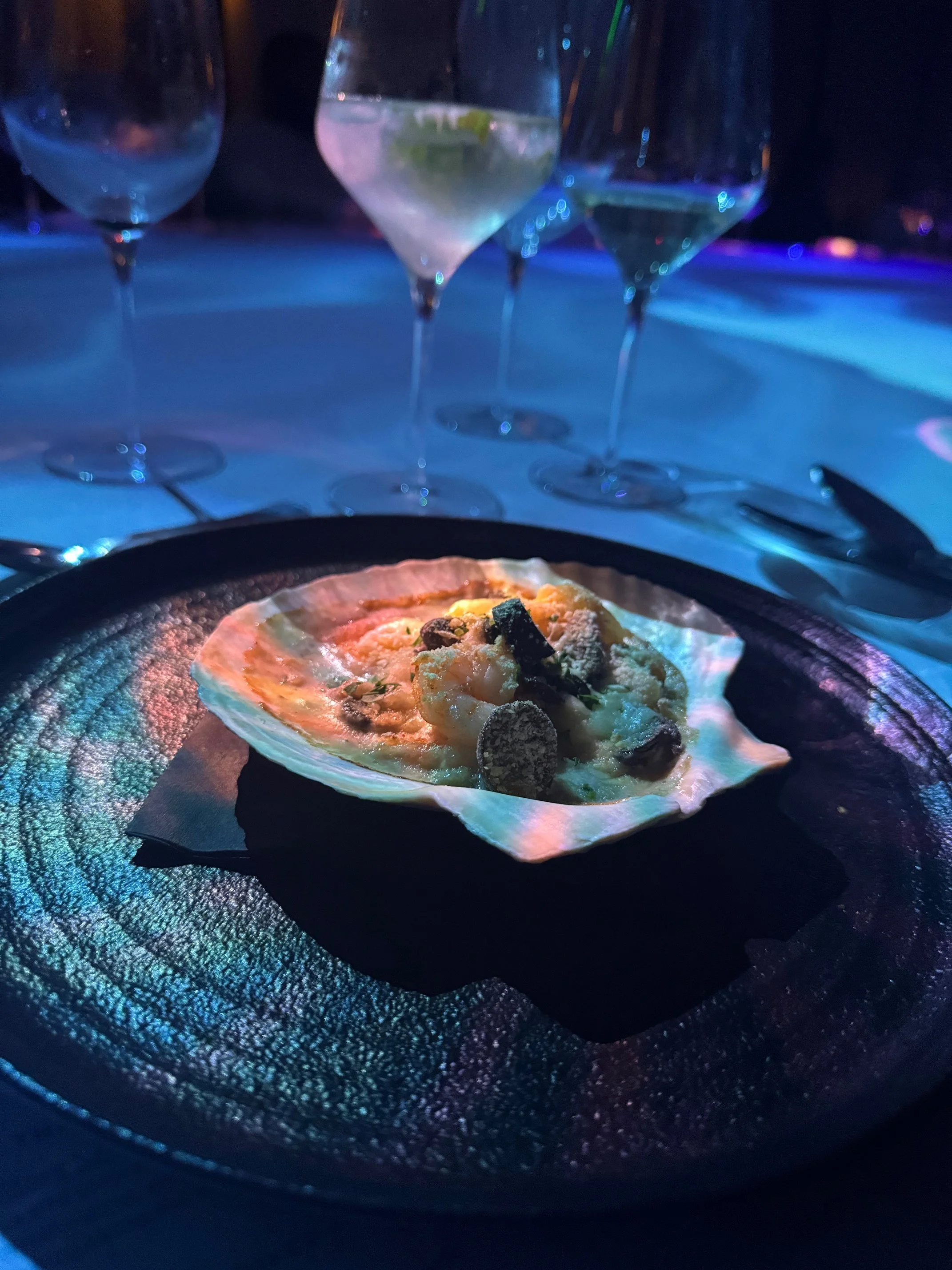 A small pizza on a black plate with toppings including cheese, vegetables, and possibly meat. The pizza is being cooked on a grill or hot stone.
In the background, there are three wine glasses filled with white wine, set on a table, with colorful lig