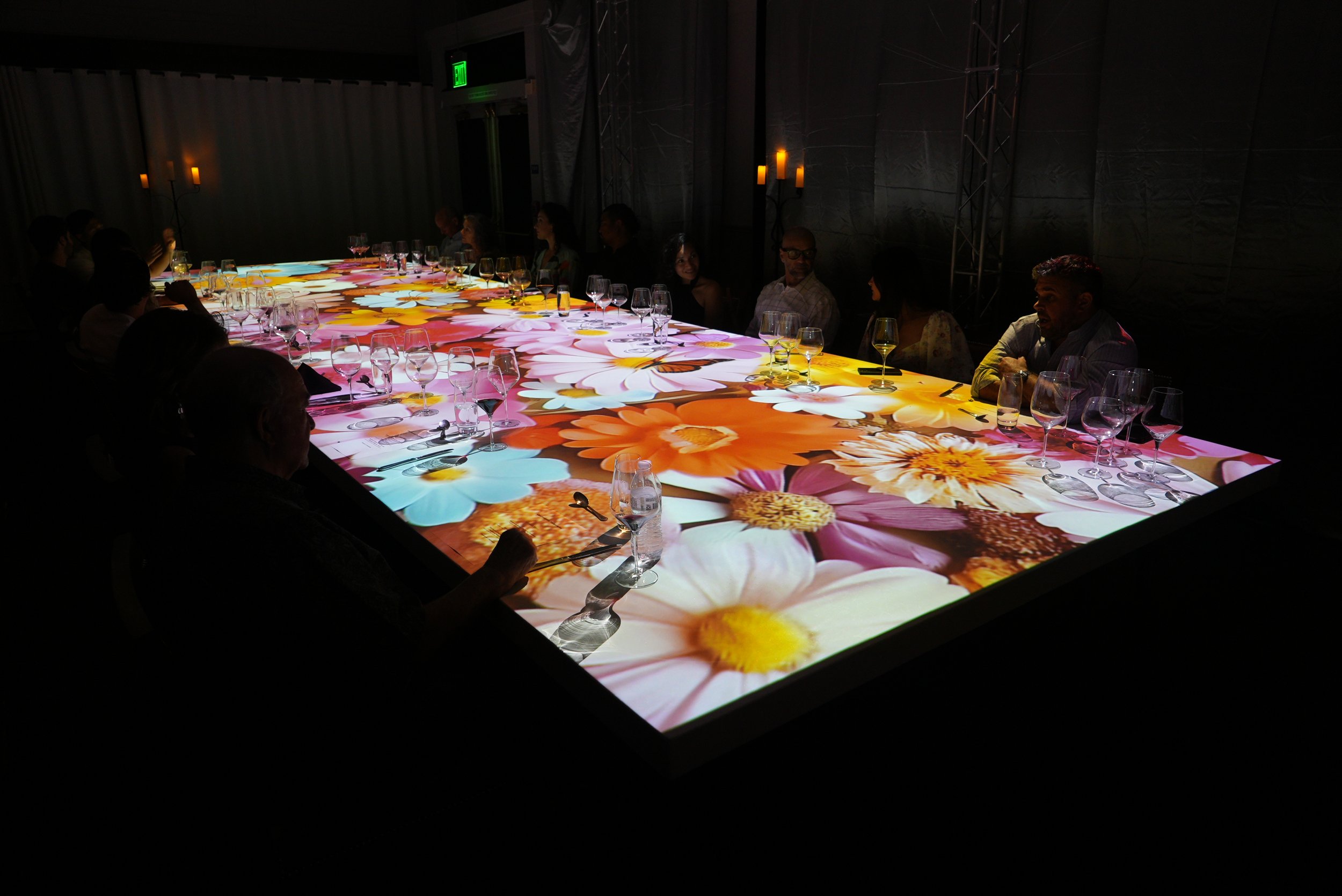 A dark room featuring a long table with a vibrant floral projection displaying large pink, orange, yellow, and white flowers. Several people are seated around the table, and some are visible on the far side. The table is set with wine glasses and din