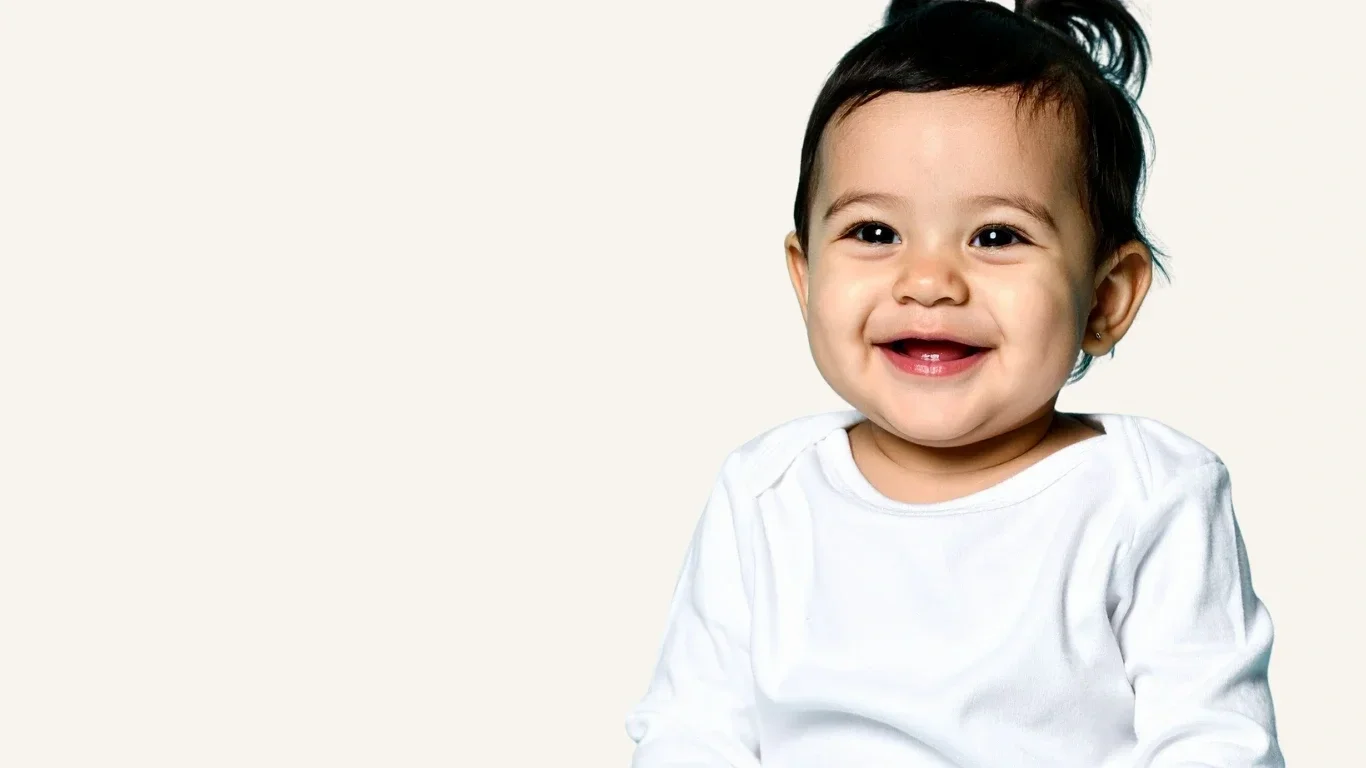 Smiling young child with dark hair and a white shirt, sitting against a light background.
