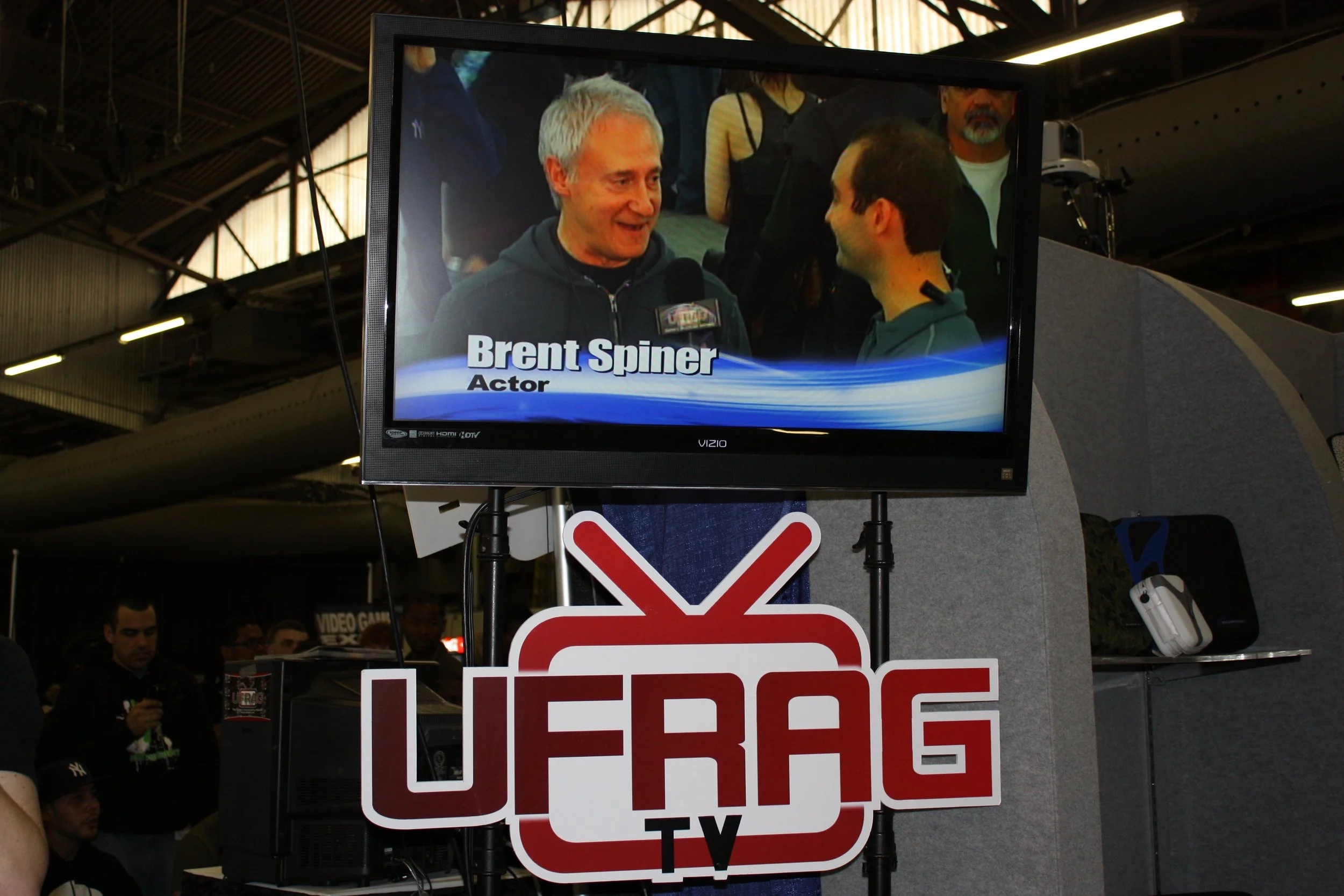 A television screen showing a man being interviewed, identified as Brent Spiner, with the title "Actor". The screen is mounted above a sign that reads "UFRAG TV". The setting appears to be at an event or convention.