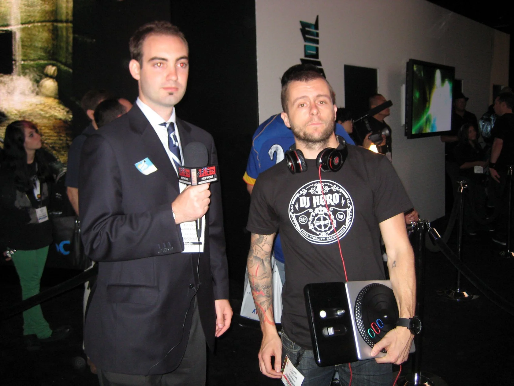 This image shows a man wearing a suit and holding a microphone, standing next to another man with headphones around his neck, holding a laptop, in a busy exhibition or conference setting.