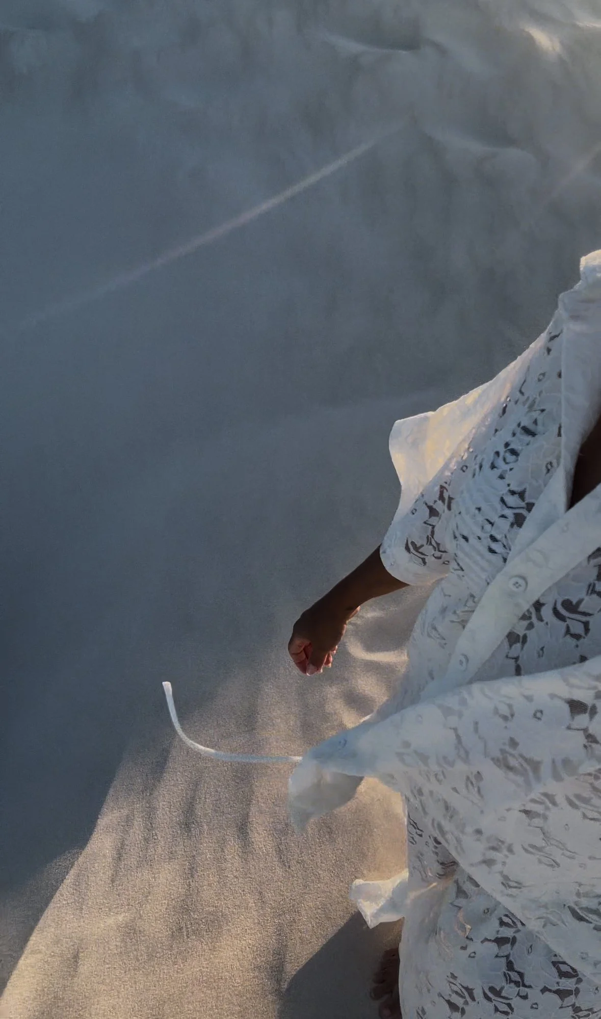 A person in white lace clothing walking along a sandy beach, with ocean water and waves in the background, and the sun casting a warm glow.