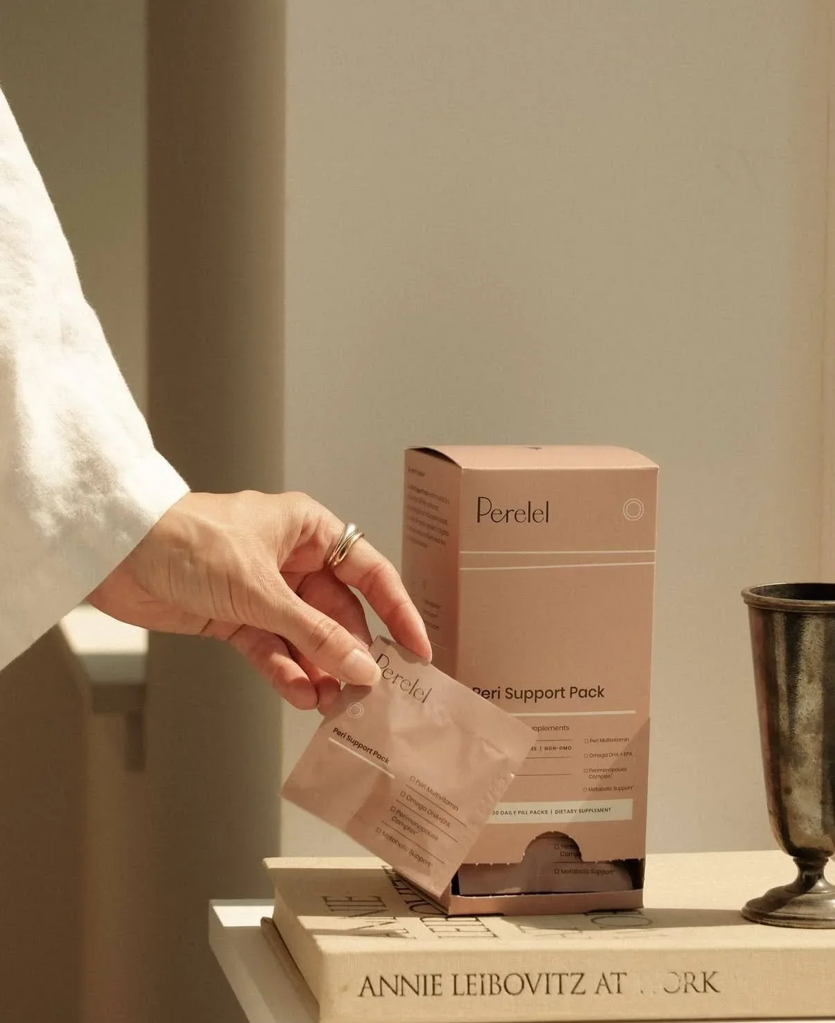 A hand with a ring is removing a pink packet labeled 'Perel Support Pack' from a box with the same label, placed on a stack of books and next to a metal container.