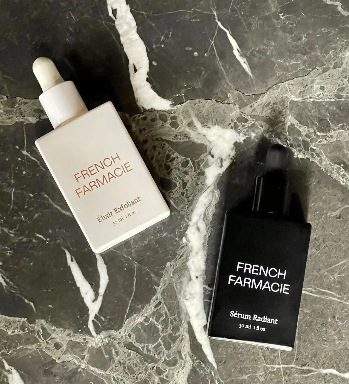 Two skincare products from French Farmacie on a marble surface. One is a white bottle labeled 'Élixir Exfoliant,' and the other is a black bottle labeled 'Sérum Radiant'.
