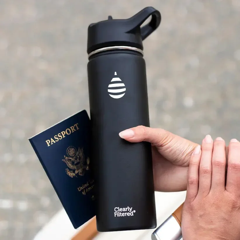 A person's hand holding a black reusable water bottle with a white logo and the text "Clearly Filtered". The person is also holding a U.S. passport, and the background is a blurry outdoor surface.