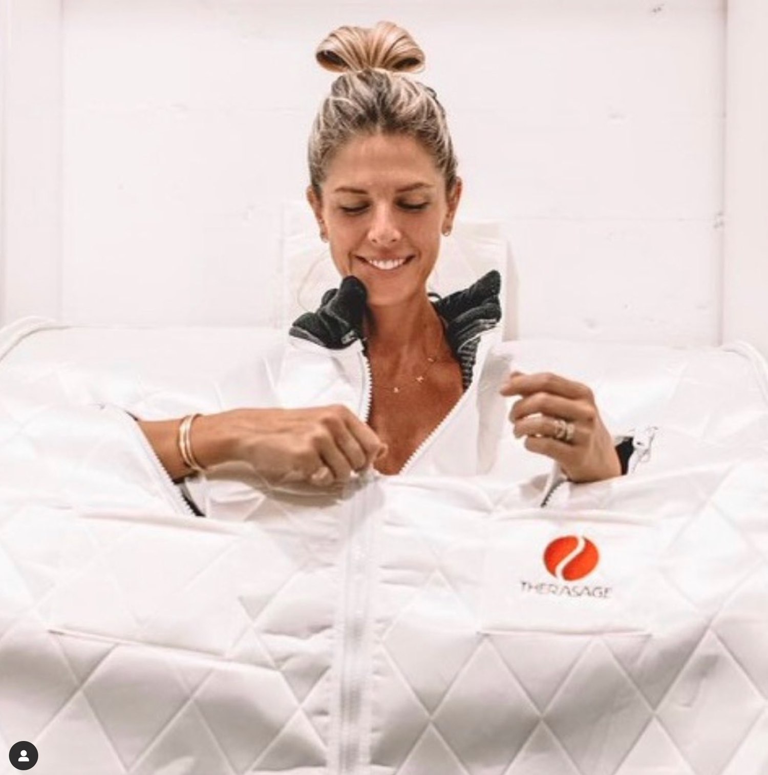 A woman smiling in a white jacket with the TherAsage logo, sitting on a bed with a white quilted cover, holding a small card and a peel-off from the cover.