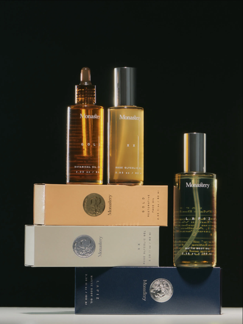 Three bottles of Monastery skincare products and their respective boxes arranged on a black background. The bottles are rectangular with metallic caps, containing different oils or serums, with one dropper bottle in the center. The boxes are stacked below the bottles, with labels indicating gold, rose, and another product.