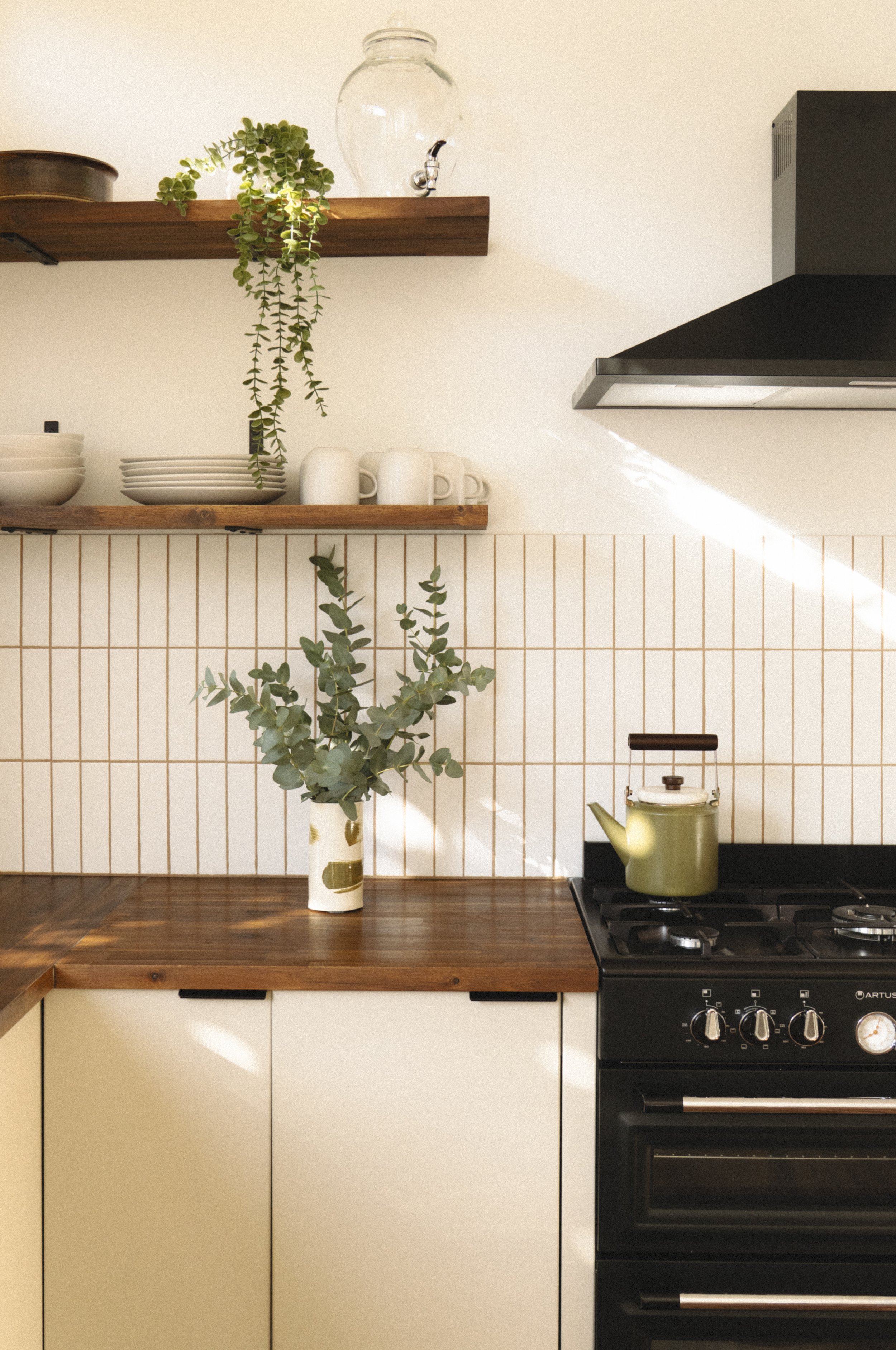 Kitchen with white tile backsplash, wooden countertop, and black stove. Open wooden shelves with dishes, cups, and plants. Green plants in the foreground and on the shelves, with sunlight streaming in.