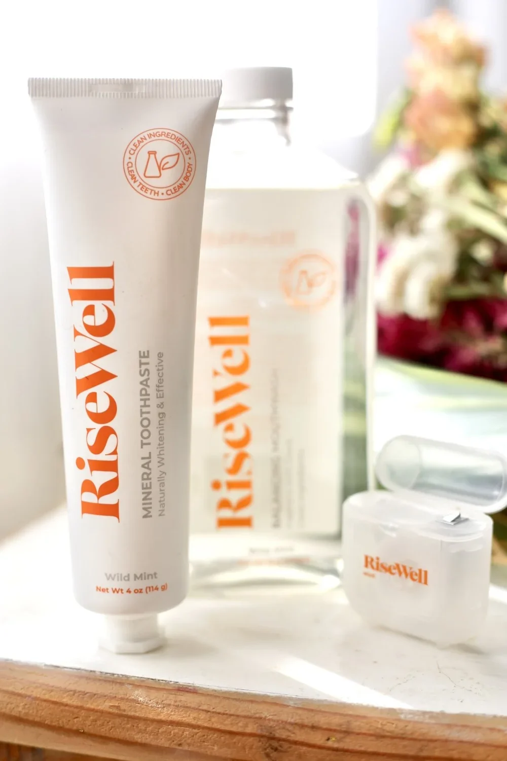 A tube of Risewell mineral toothpaste with natural whitening and effective ingredients in Wild Mint flavor, placed on a white surface with a Risewell bottle and a small container nearby, and some flowers in the background.