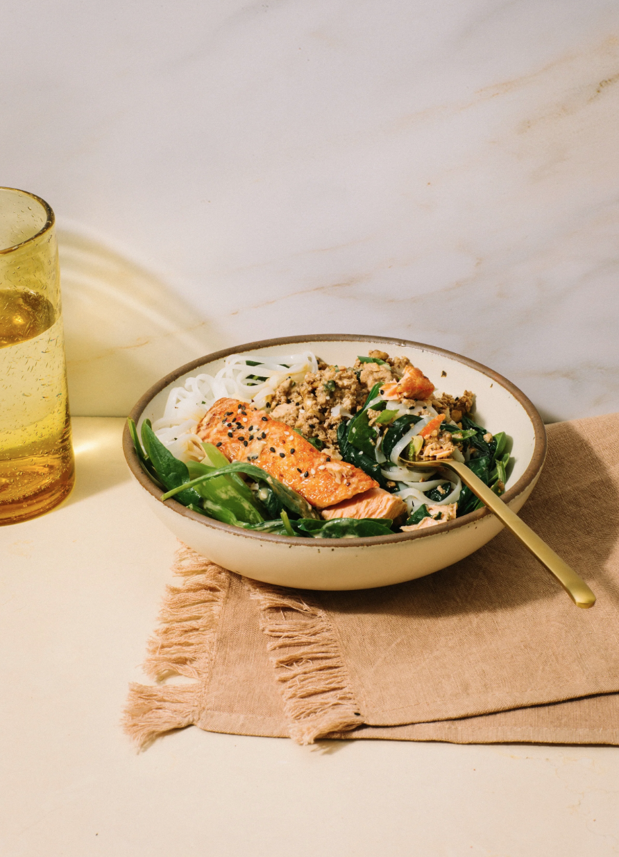 Bowl of Asian-inspired noodles with grilled salmon and vegetables, accompanied by a glass of iced tea on a beige cloth napkin on a white surface.