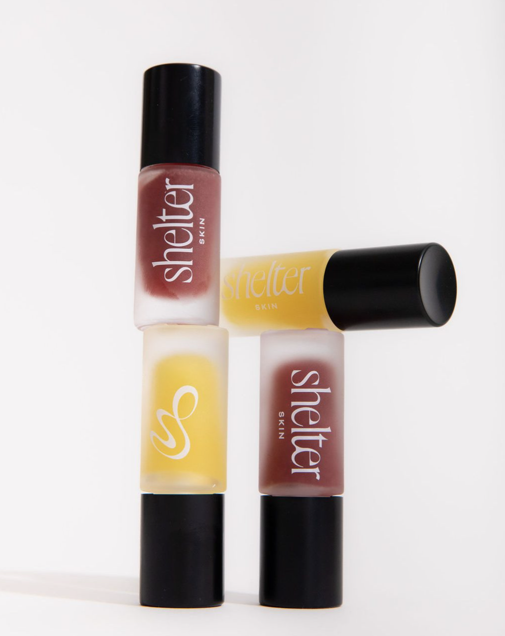 Four bottles of SHELTER SKIN lip gloss arranged in a cross shape on a white background.