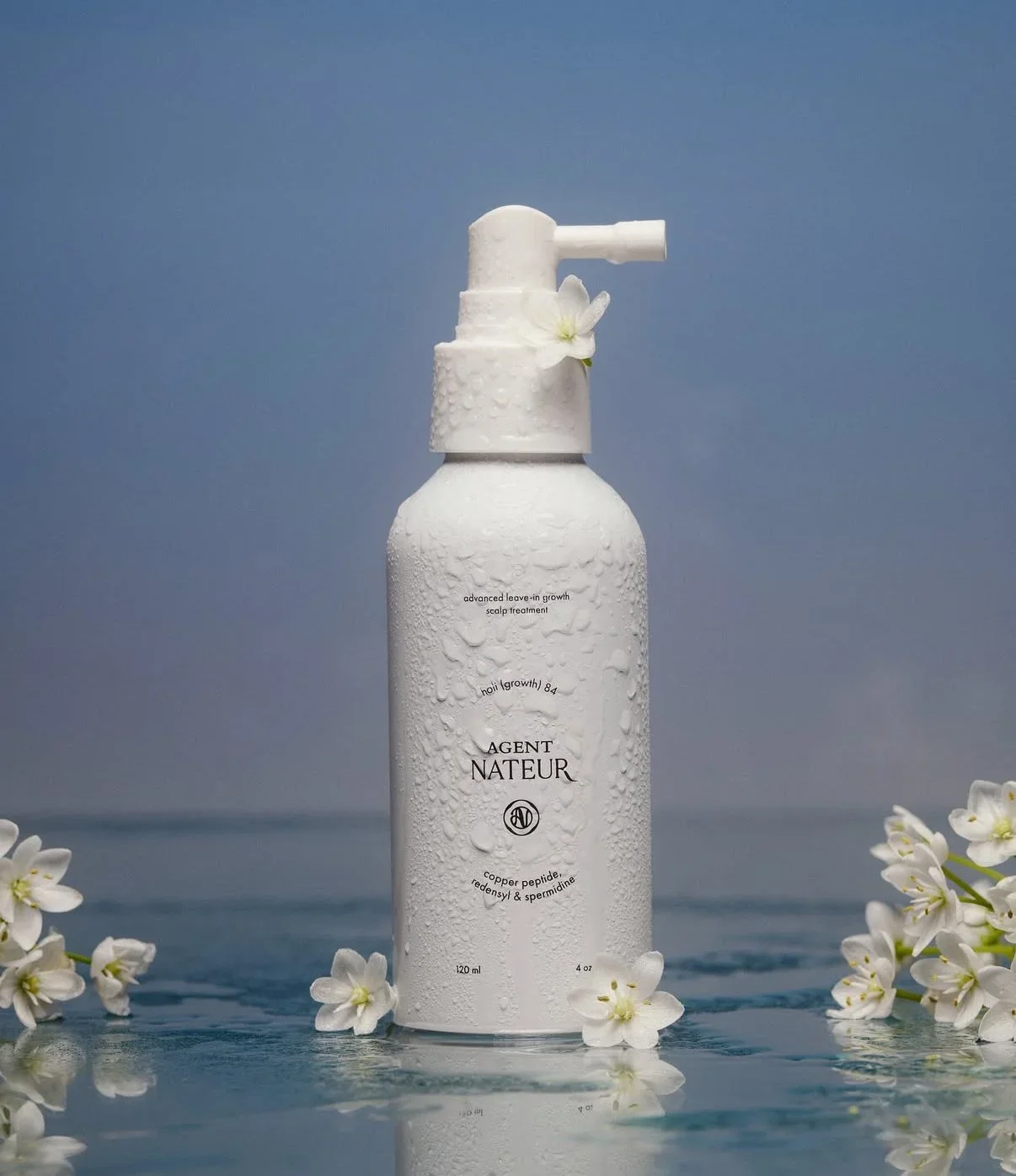 A white spray bottle labeled 'Agent Nature' with droplets of water on it, surrounded by white flowers on a reflective surface against a light blue background.
