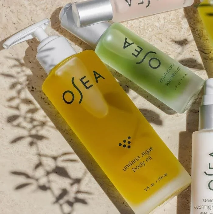 Several bottles of skincare products, including yellow, green, white, and yellowish-green bottles, are placed on a sandy surface with shadows of leaves cast on the ground. The bottles are branded 'OSEA'.