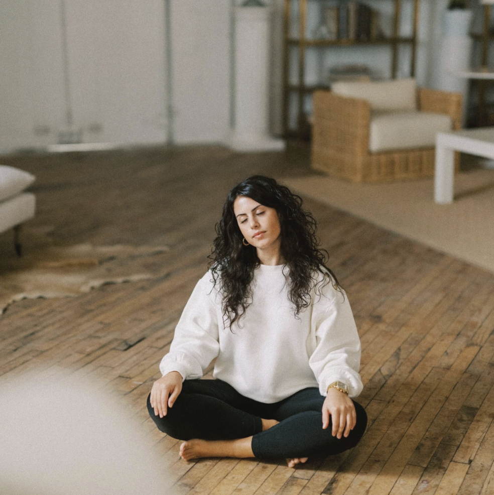 A woman with curly dark hair sitting cross-legged on a wooden floor in a living room, eyes closed, appearing to meditate. Anthea Dagiantis, Aeonian Wellness, Gut Health Coach in New Jersey.