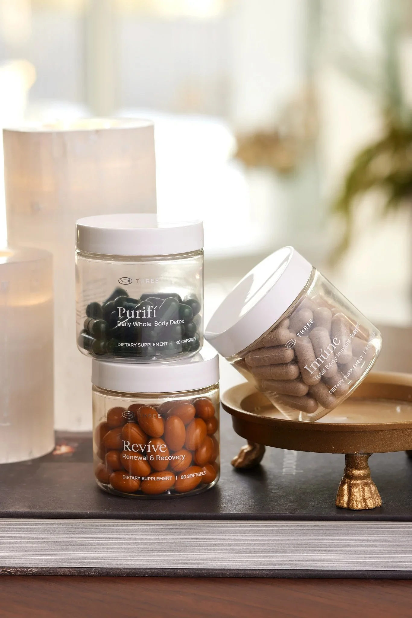 Stacked clear containers with supplement pills on a dark-colored surface, with a small decorative gold-legged tray to the right.