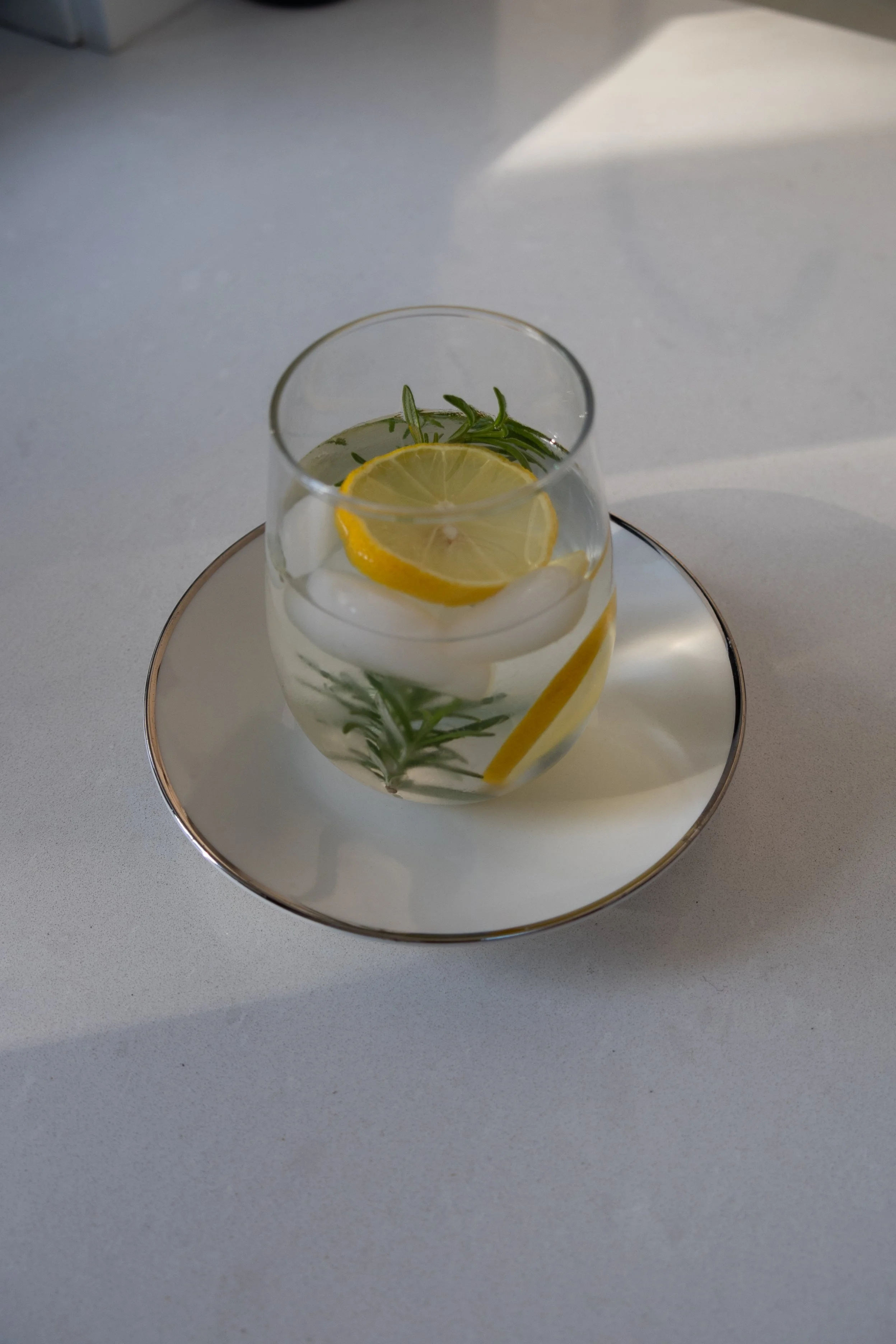 A glass of water with slices of lemon and sprigs of rosemary, placed on a saucer on a white surface. Aeonian Wellness, Gut Health Coach in New Jersey.