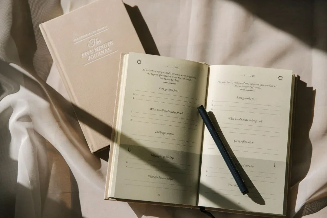 An open journal with prompts for gratitude and daily reflections on a white table, with a black pen on the right page, and a closed book titled "The Five Minute Journal" on the left side.