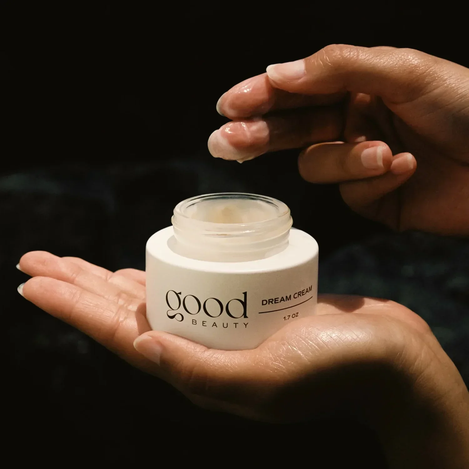 Close-up of a person's hand holding an open jar of cream labeled 'good beauty dream cream' with black background.