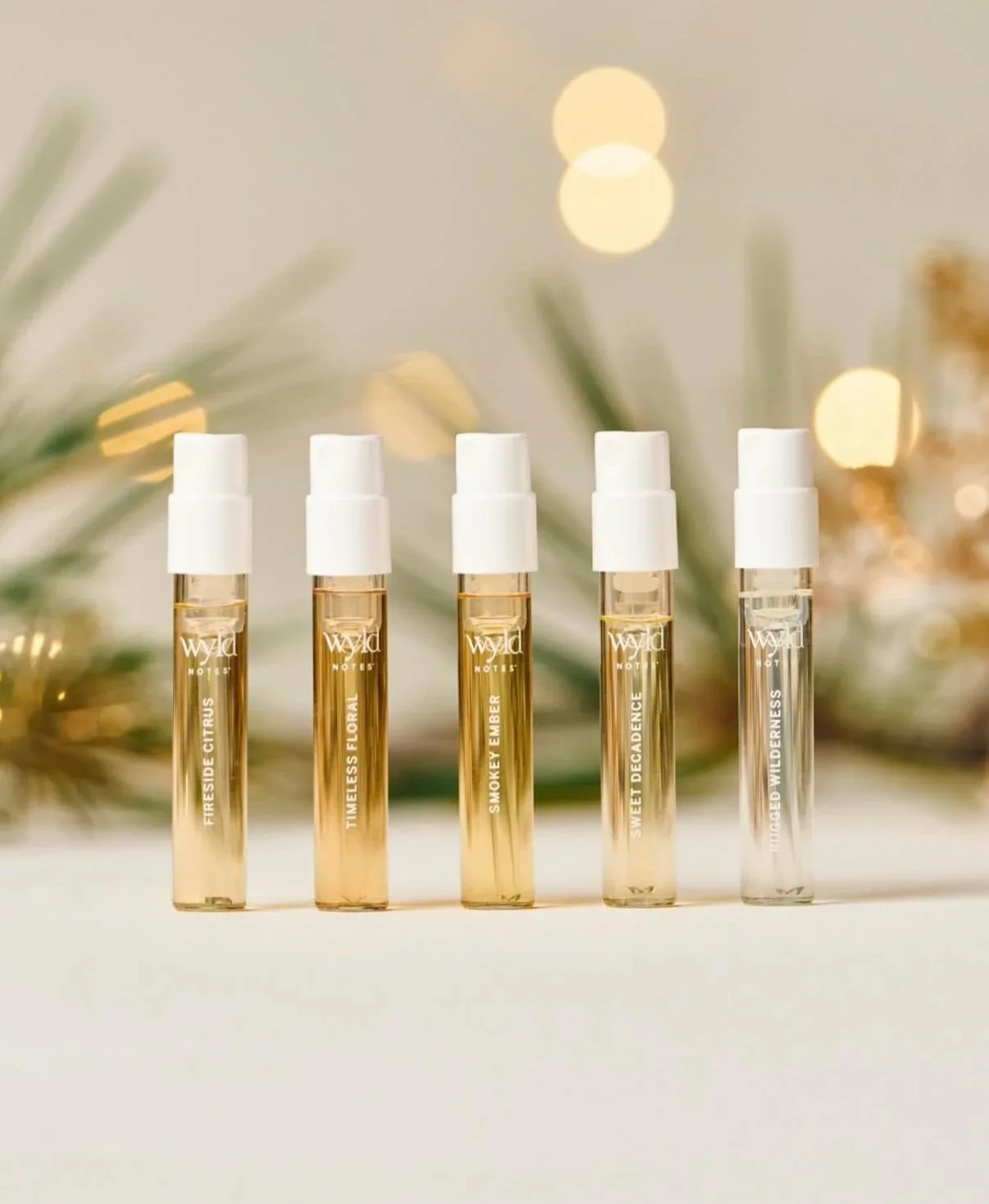 Five small perfume spray bottles with white caps, labeled with different fragrance names, arranged in a row against a blurred background of holiday greenery and warm lights.